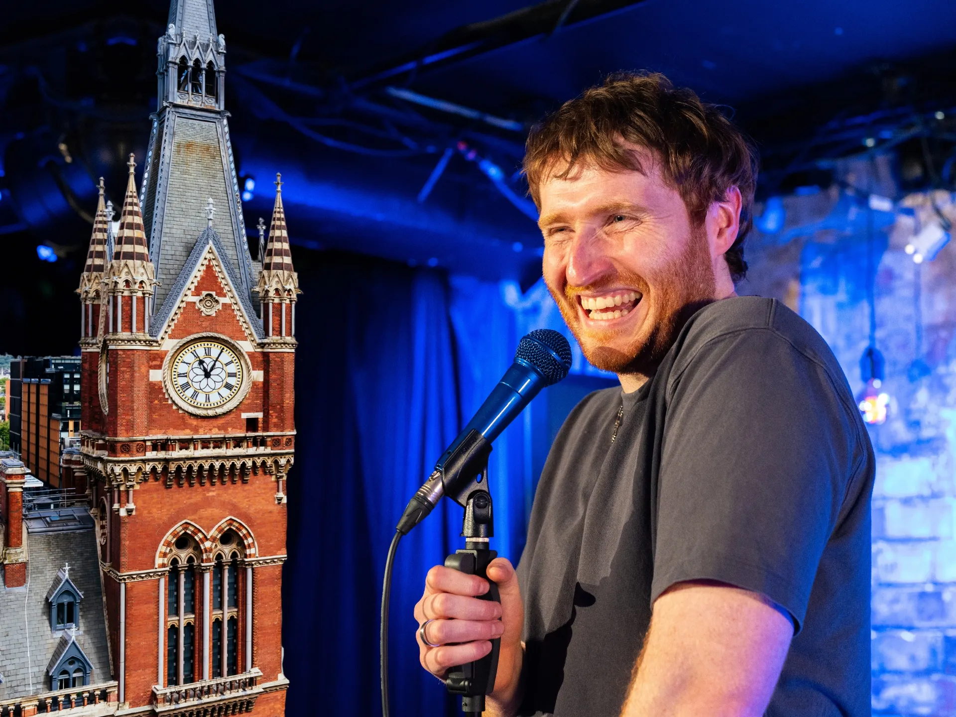 British stand-up comedian Ali Woods performing at Comedy Freaks Saturday night comedy show inside historic St Pancras Clock Tower, Kings Cross London - Victorian Gothic architecture with clock face visible alongside blue stage lighting