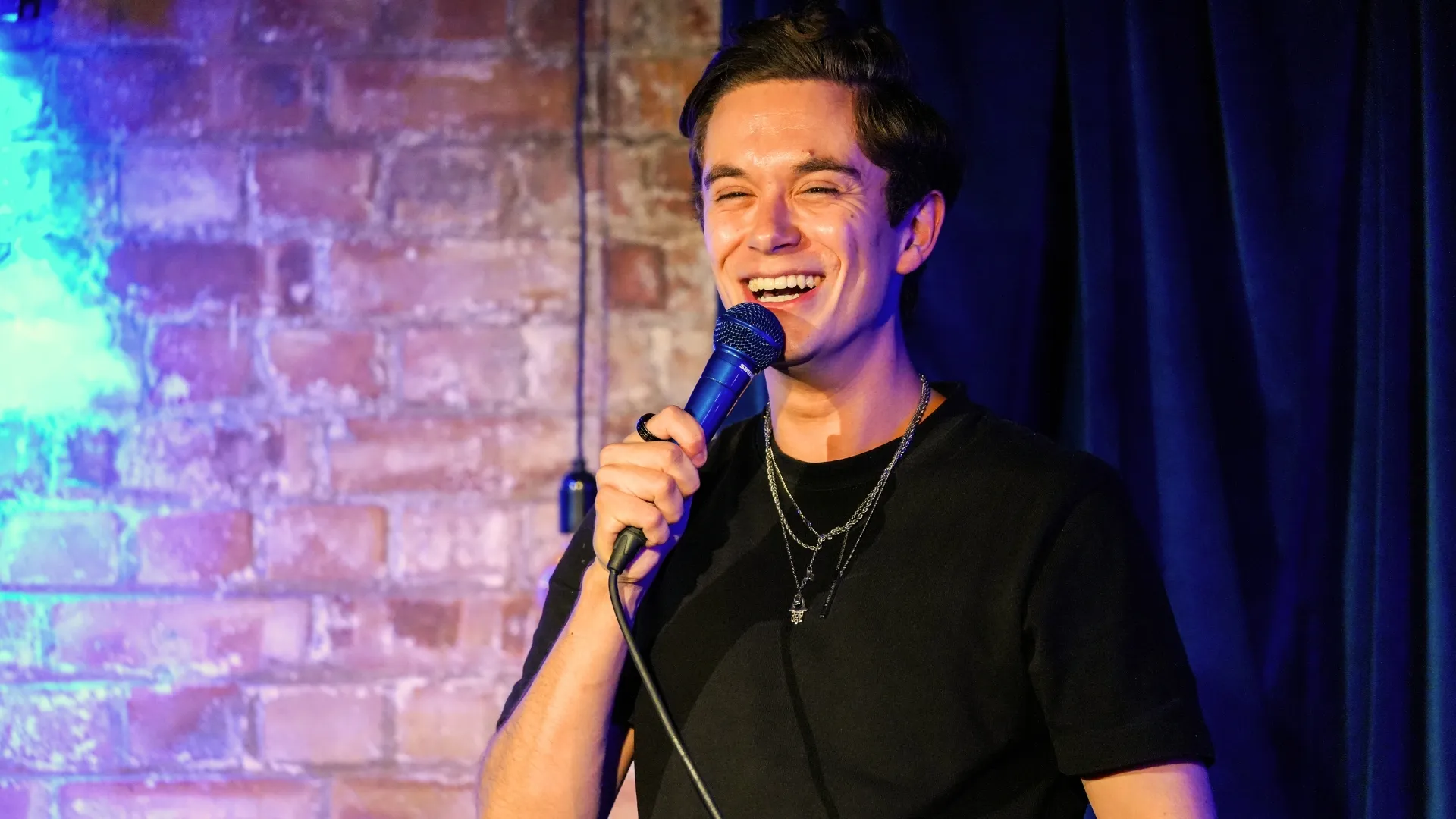 Jomi Cruz, a male comedian with dark hair, laughs while holding a microphone on stage against a brick wall with blue lighting, delivering an energetic performance at Comedy Freaks - The Most Unique Comedy Club in London.