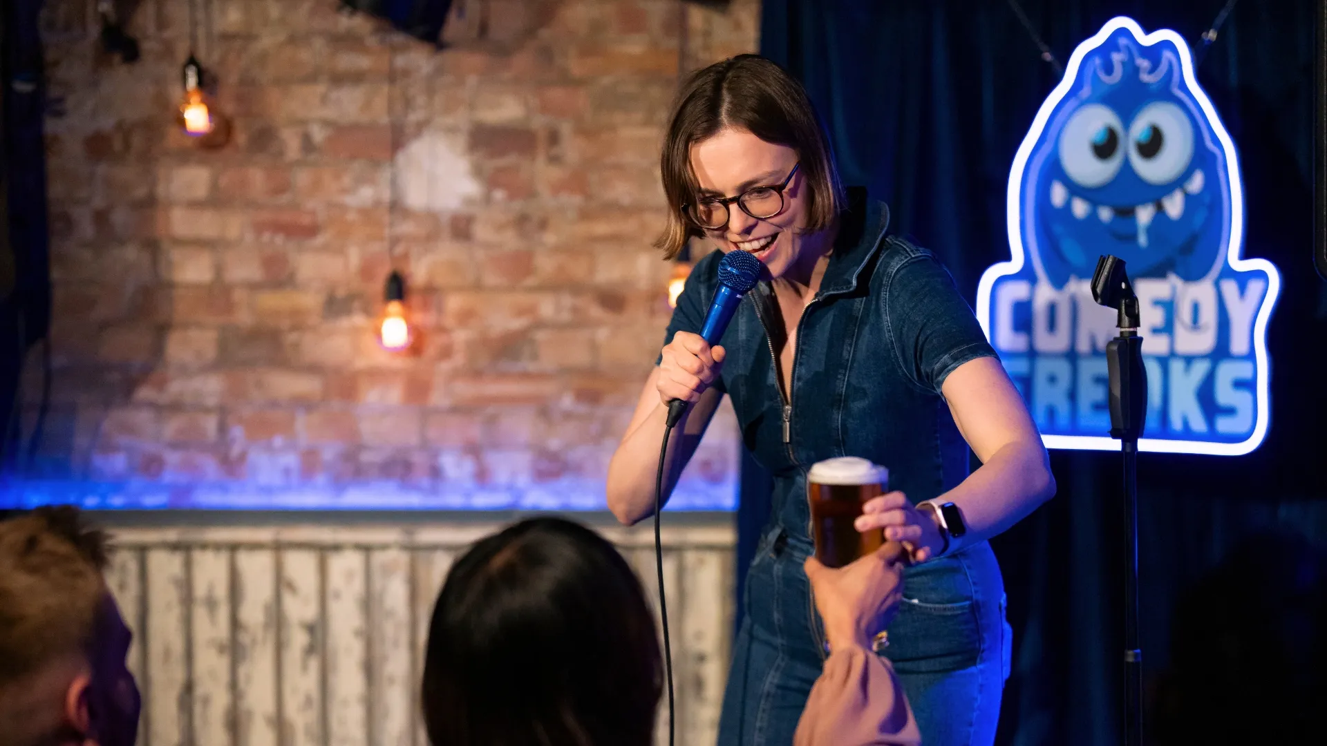 Katie Kamola interacts playfully with an audience member, holding a microphone and a drink, on stage in front of the glowing Comedy Freaks logo, showcasing the audience engagement and vibrant atmosphere of London best comedy show
