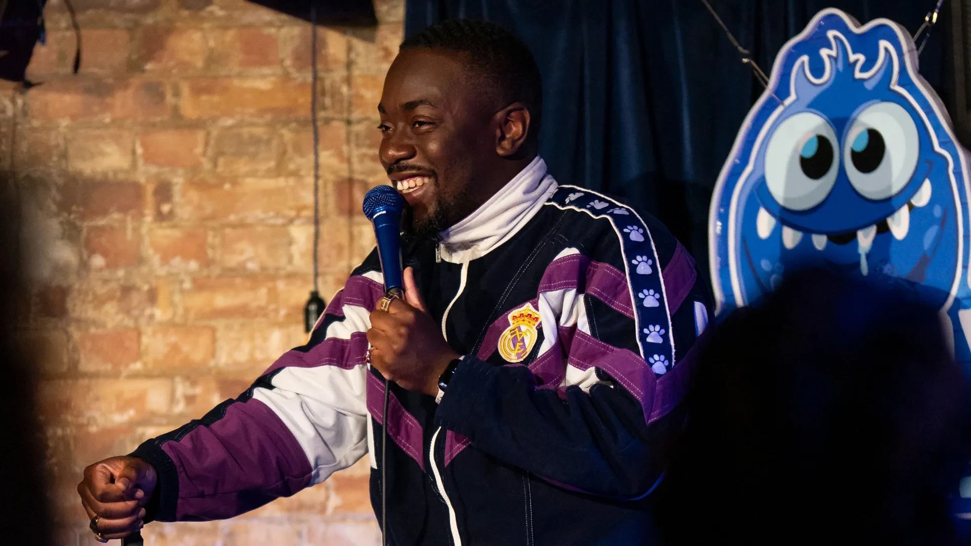 Andrew Mensah, a male black comedian, smiles while holding a microphone and gesturing with his other hand, on stage with a brick wall backdrop and the glowing blue Comedy Freaks logo, delivering energetic stand-up comedy at our London comedy venue.