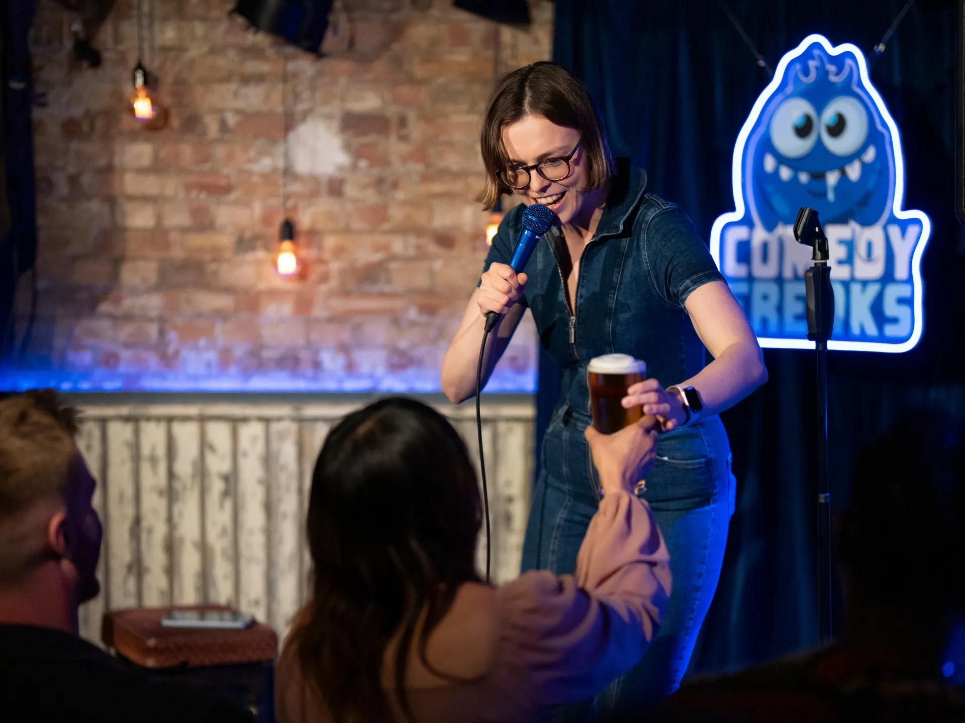 Comedian Katie Kamola delivering her set at a Comedy Freaks comedy show in London, wearing glasses and black denim jacket, casually holding a pint of beer while speaking into a microphone, with the vibrant blue Comedy Freaks monster logo and brick venue architecture behind her.