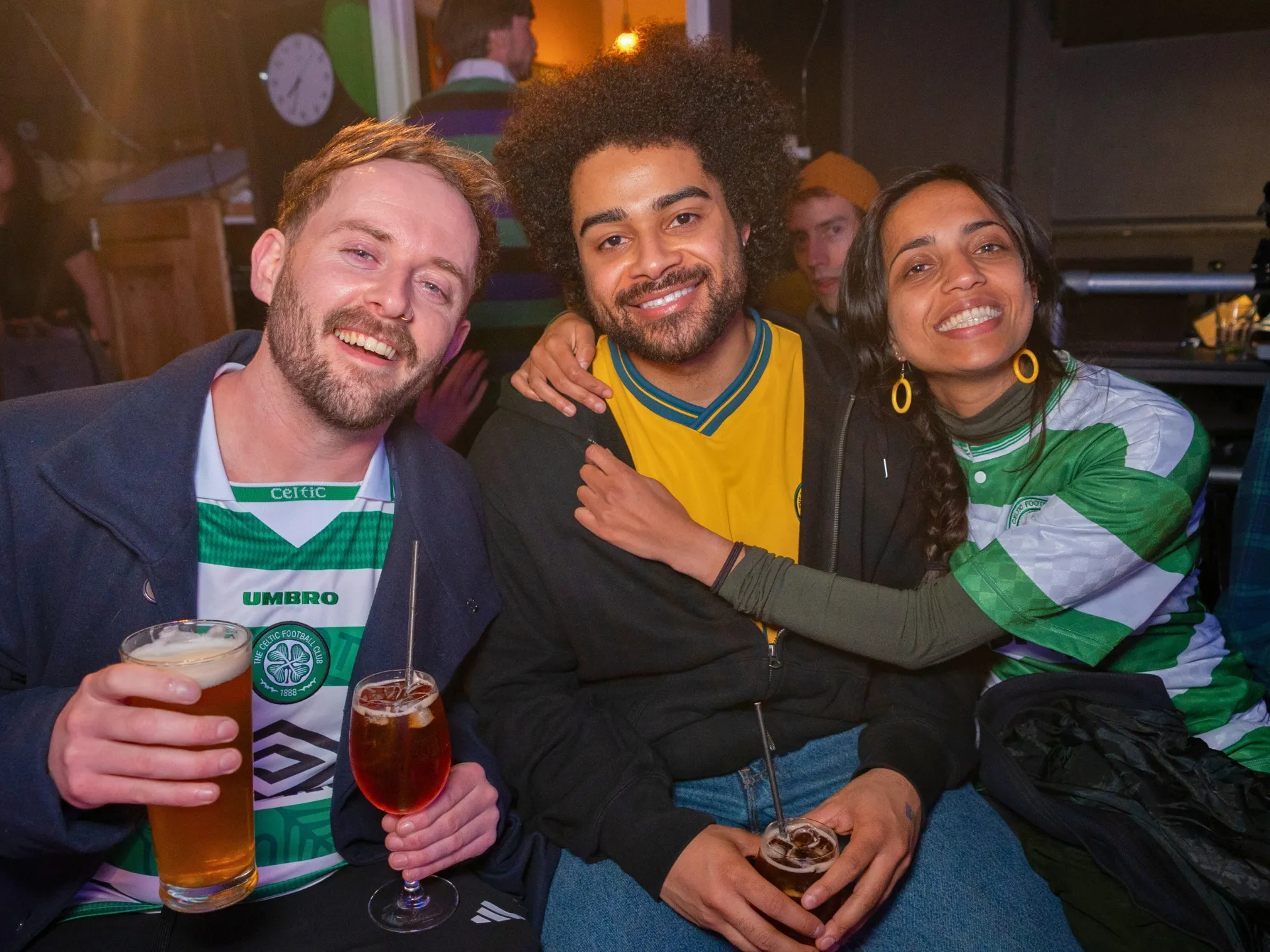 Three smiling audience members enjoying a Comedy Freaks comedy show in London, two men and one woman holding pints of beer, wearing green and white Celtic football jerseys, warm pub lighting creating a friendly atmosphere as they pose together during the interval with drinks in hand.