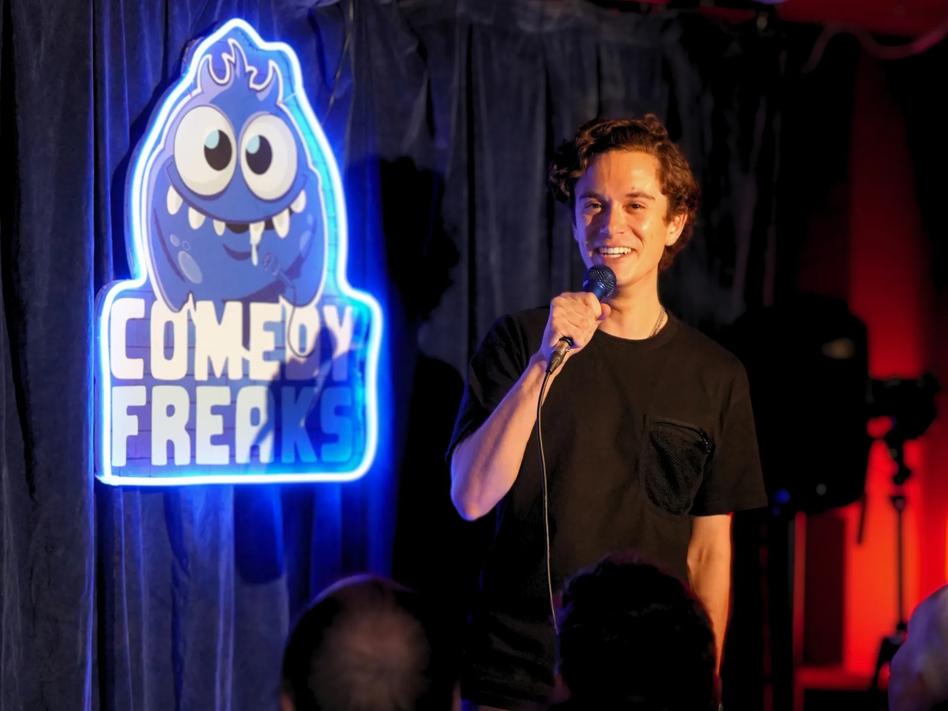 UK comedian Jomi Cruz mid-set at a London comedy show at Comedy Freaks. He has dark curly hair and relaxed expression as he speaks into the microphone, wearing a simple black tee, the iconic blue neon monster logo and red ambient lighting creating a vibrant stage backdrop.