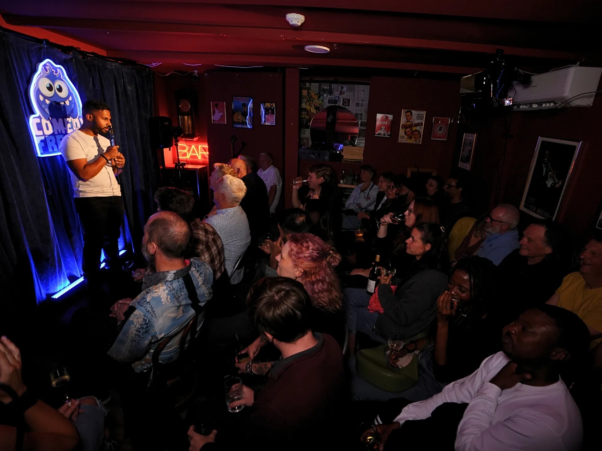 London comedian Raj Sivade commanding the stage at Comedy Freaks London Comedy show, the best Camden comedy venue. The audience members seated with drinks watching intently beneath the glowing blue neon monster logo, intimate space buzzing with energy, red BAR sign and warm lighting throughout.