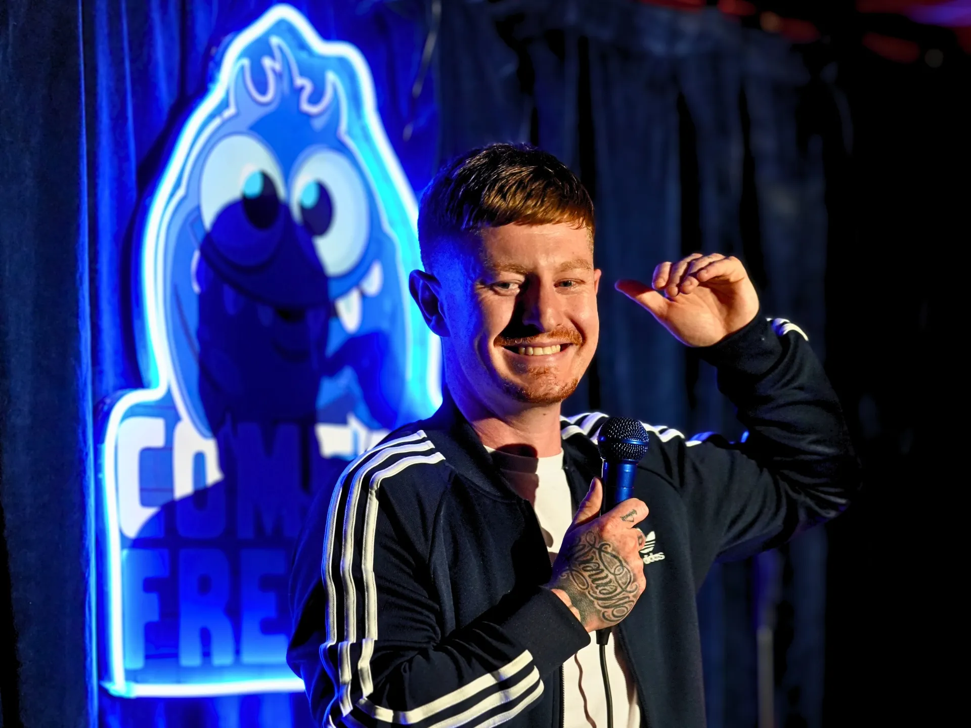  British comedian Tom Nestor on stage at a Comedy Freaks comedy show London event, wearing a sporty black Adidas track top, short ginger hair catching the blue stage lighting, holding the microphone close while smiling, the iconic neon Comedy Freaks logo glowing brightly behind him.
