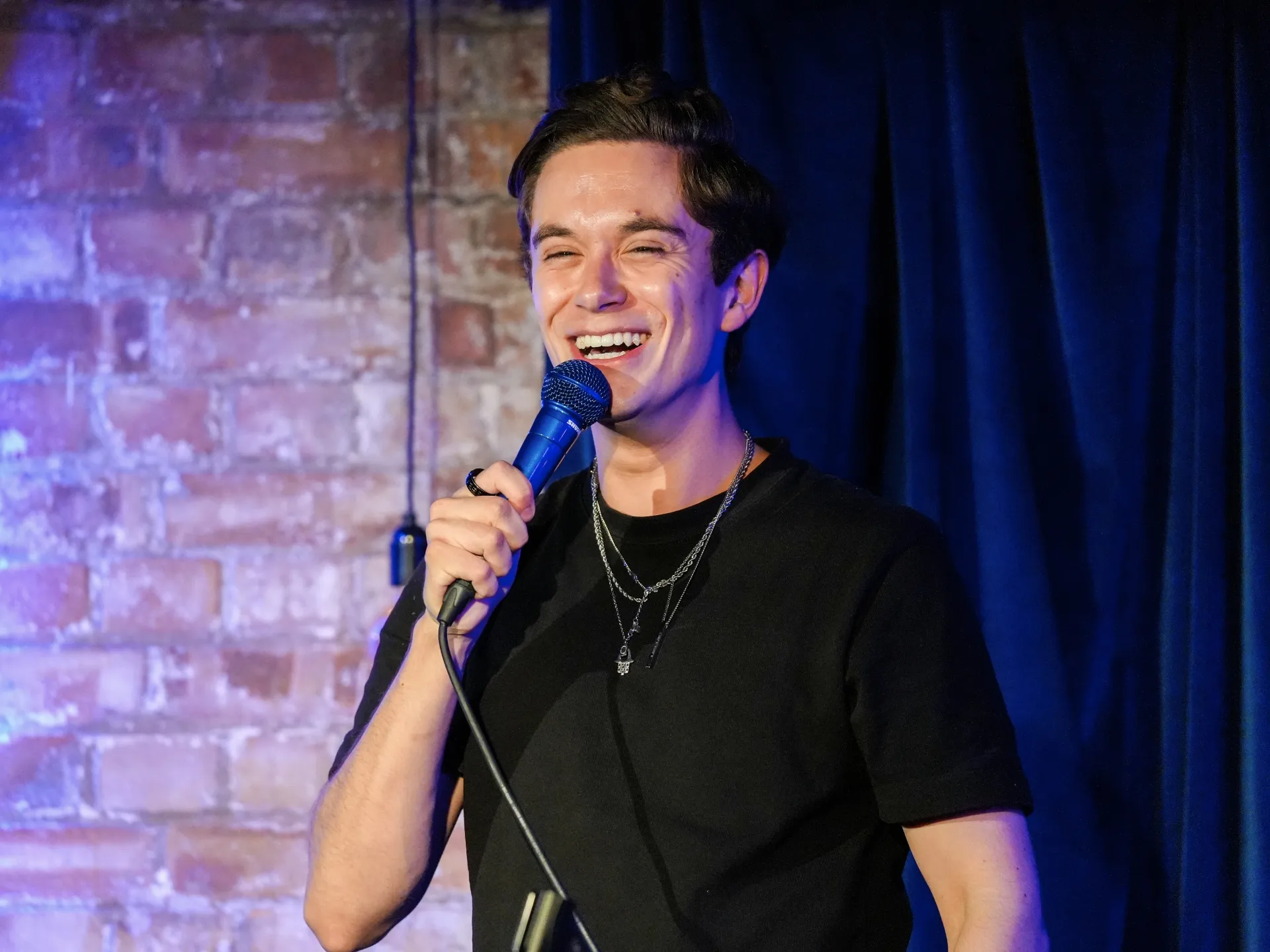 Comedian Jomi Cruz performing stand-up comedy in London at a Comedy Freaks London comedy club show, wearing a black t-shirt and silver chain necklace, holding the microphone with a warm genuine smile, exposed brick wall and soft amber stage lighting creating an intimate comedy show atmosphere behind him.