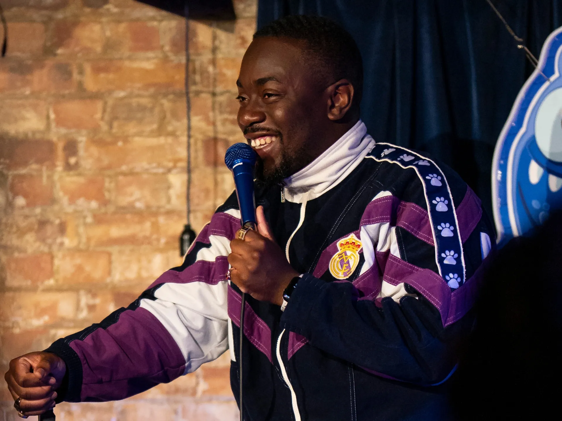  British comedian Andrew Mensah performing stand-up comedy at a Comedy Freaks London comedy show, wearing a colourful pink and navy bomber jacket over a white hoodie, big animated smile as he holds the mic mid-joke at this comedy show in London, the glowing blue neon logo and brick wall behind.