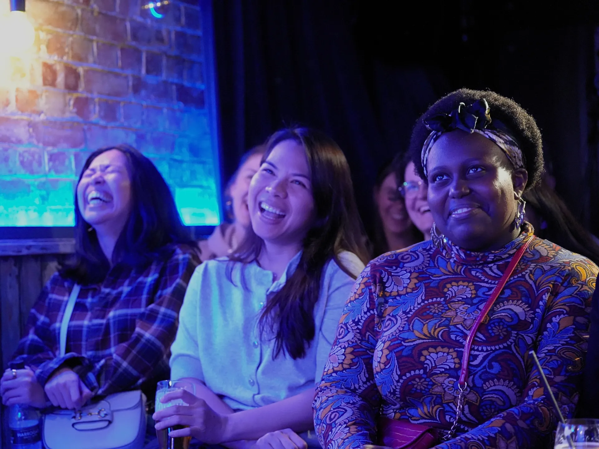 Three women in the audience laughing at Comedy Freaks comedy club show, enjoying one of the best comedy shows in London, genuine warm smiles as they watch stand-up comedy together, one in a blue checked shirt, soft blue neon lighting creating a cosy atmosphere in the background.