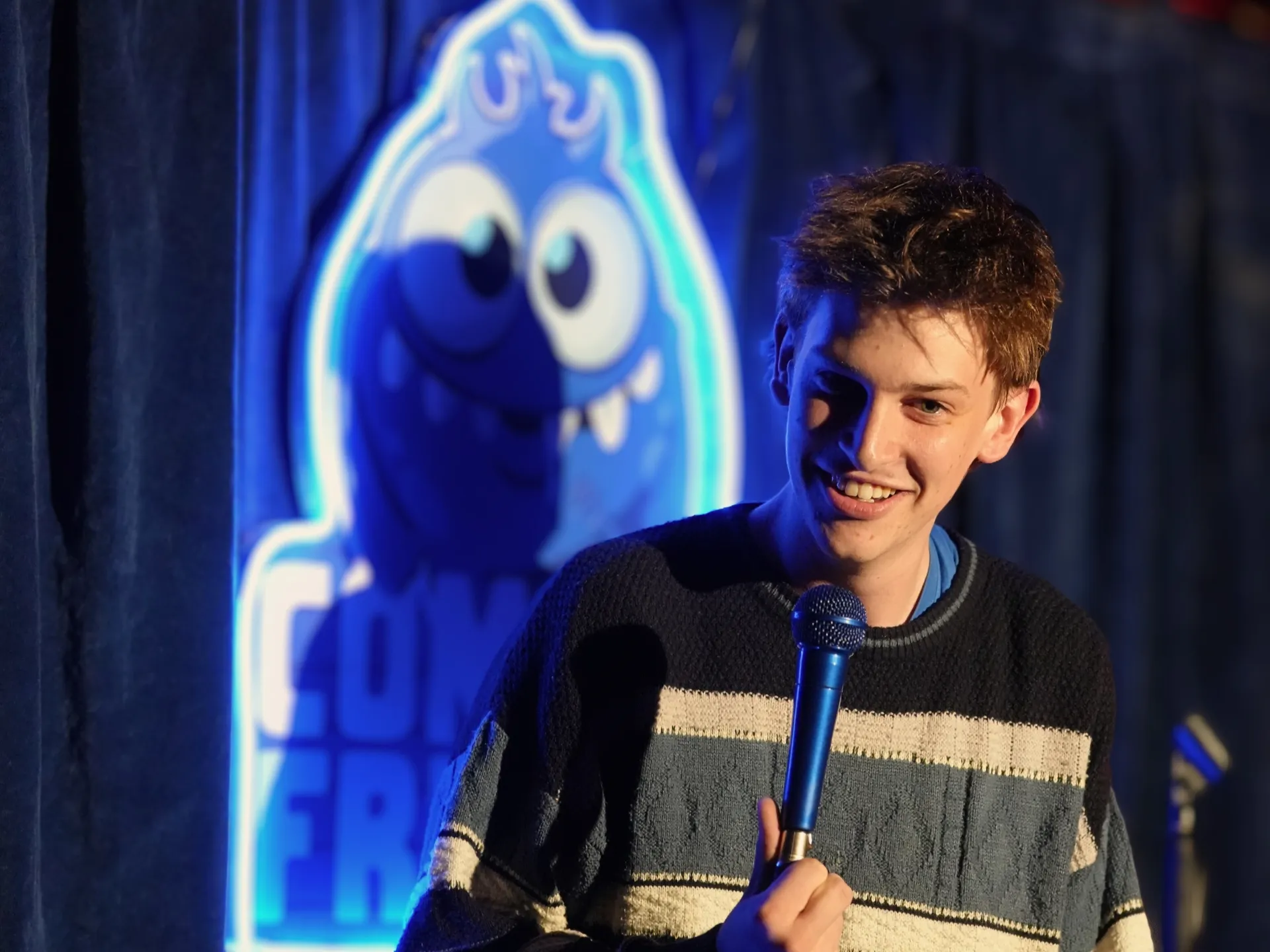 British comedian Jon Gibbons on stage at Comedy Freaks, one of the best things to do in London for live comedy shows, wearing a navy striped jumper with tousled brown hair, youthful energy at this popular comedy club as he grips the mic, the blue neon logo illuminating the backdrop.