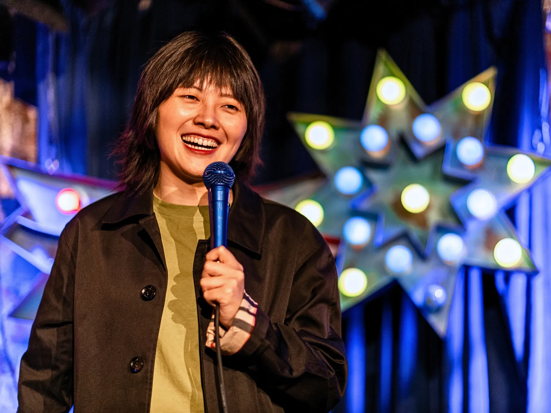 British comedian Blank Peng performing stand-up comedy at a Comedy Freaks comedy show, wearing a brown coat over an olive green top, dark hair with fringe, holding the microphone with a bright warm smile at this comedy show in London, an illuminated light decoration glowing behind her.