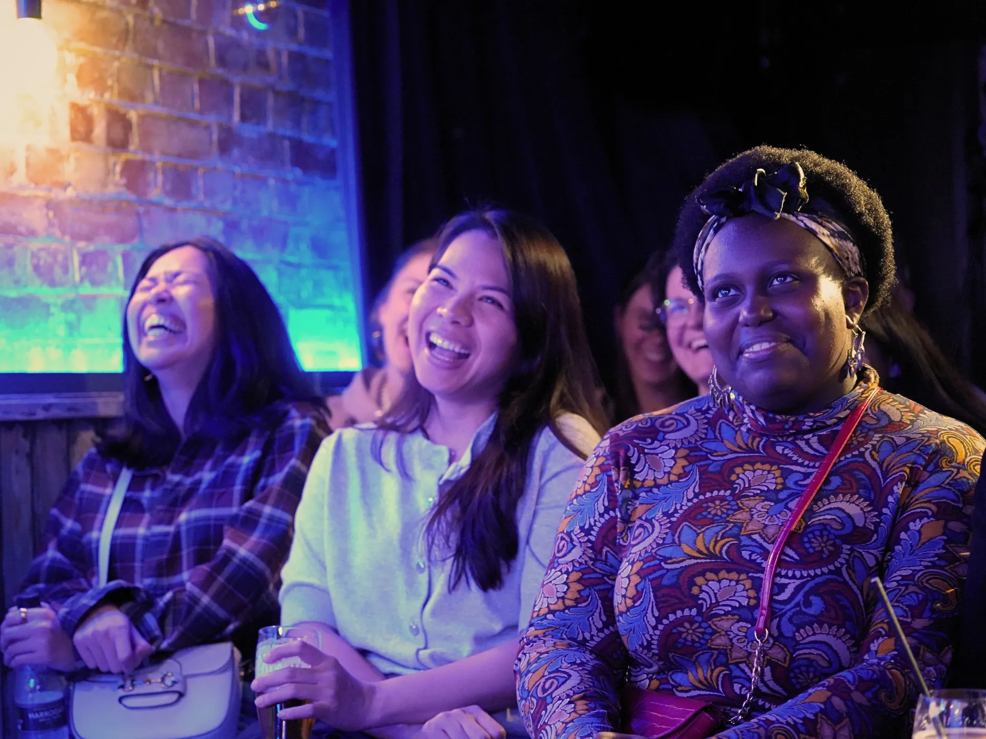 Three women in the audience laughing at Comedy Freaks comedy club show, enjoying one of the best comedy shows in London, genuine warm smiles as they watch stand-up comedy together, one in a blue checked shirt, soft blue neon lighting creating a cosy atmosphere in the background.