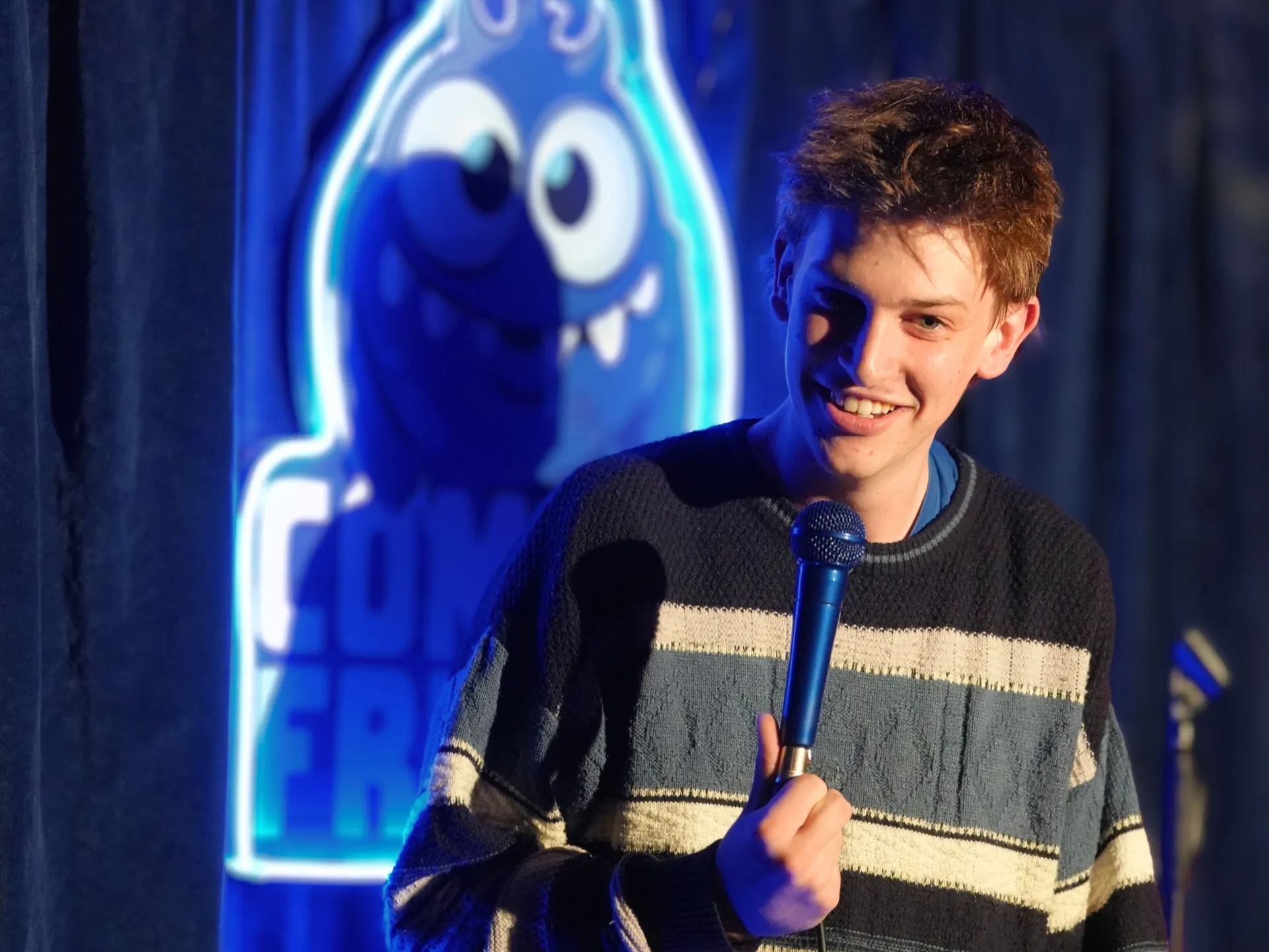 British comedian Jon Gibbons on stage at Comedy Freaks, one of the best things to do in London for live comedy shows, wearing a navy striped jumper with tousled brown hair, youthful energy at this popular comedy club as he grips the mic, the blue neon logo illuminating the backdrop.