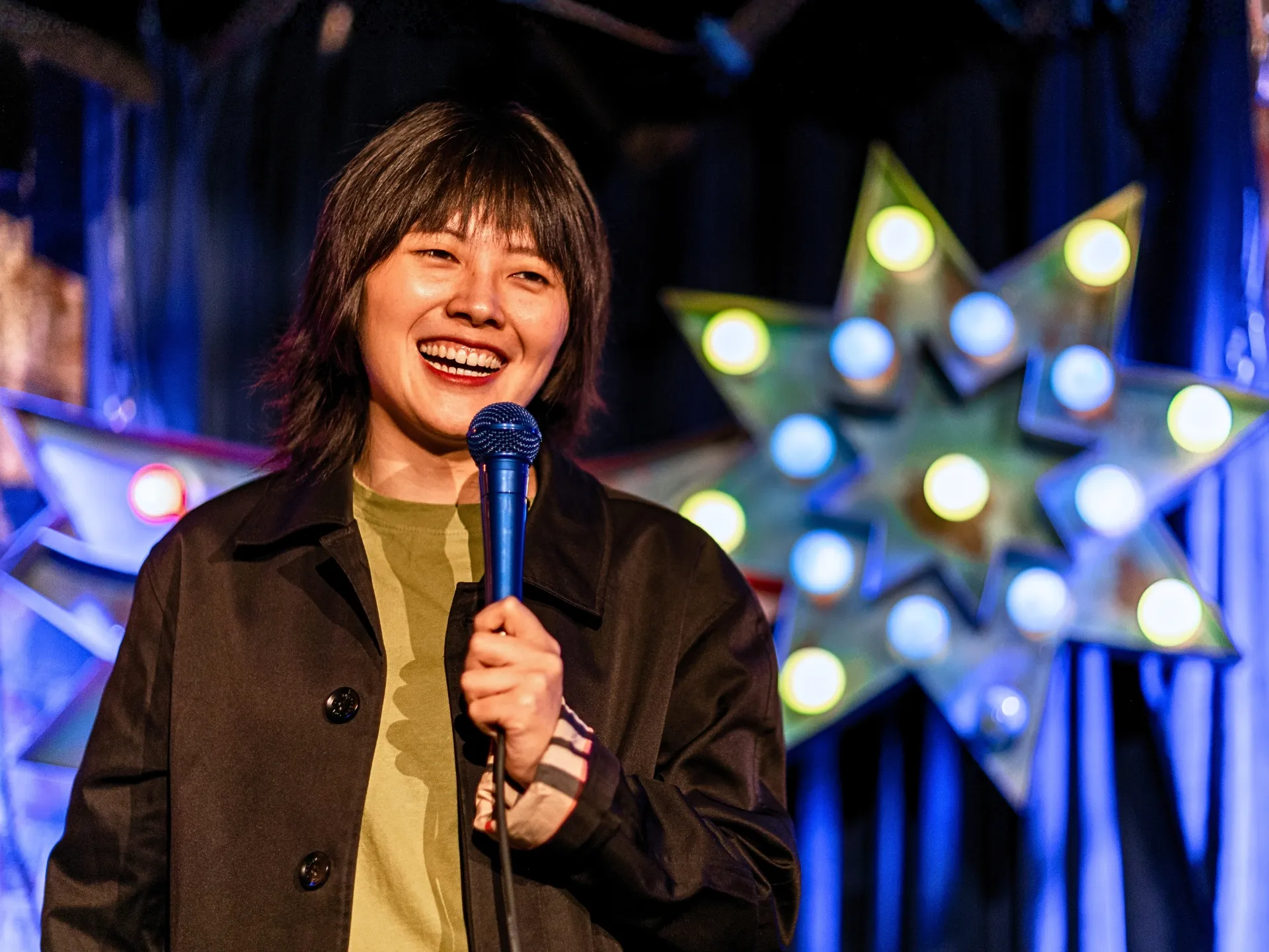 British comedian Blank Peng performing stand-up comedy at a Comedy Freaks comedy show, wearing a brown coat over an olive green top, dark hair with fringe, holding the microphone with a bright warm smile at this comedy show in London, an illuminated light decoration glowing behind her.