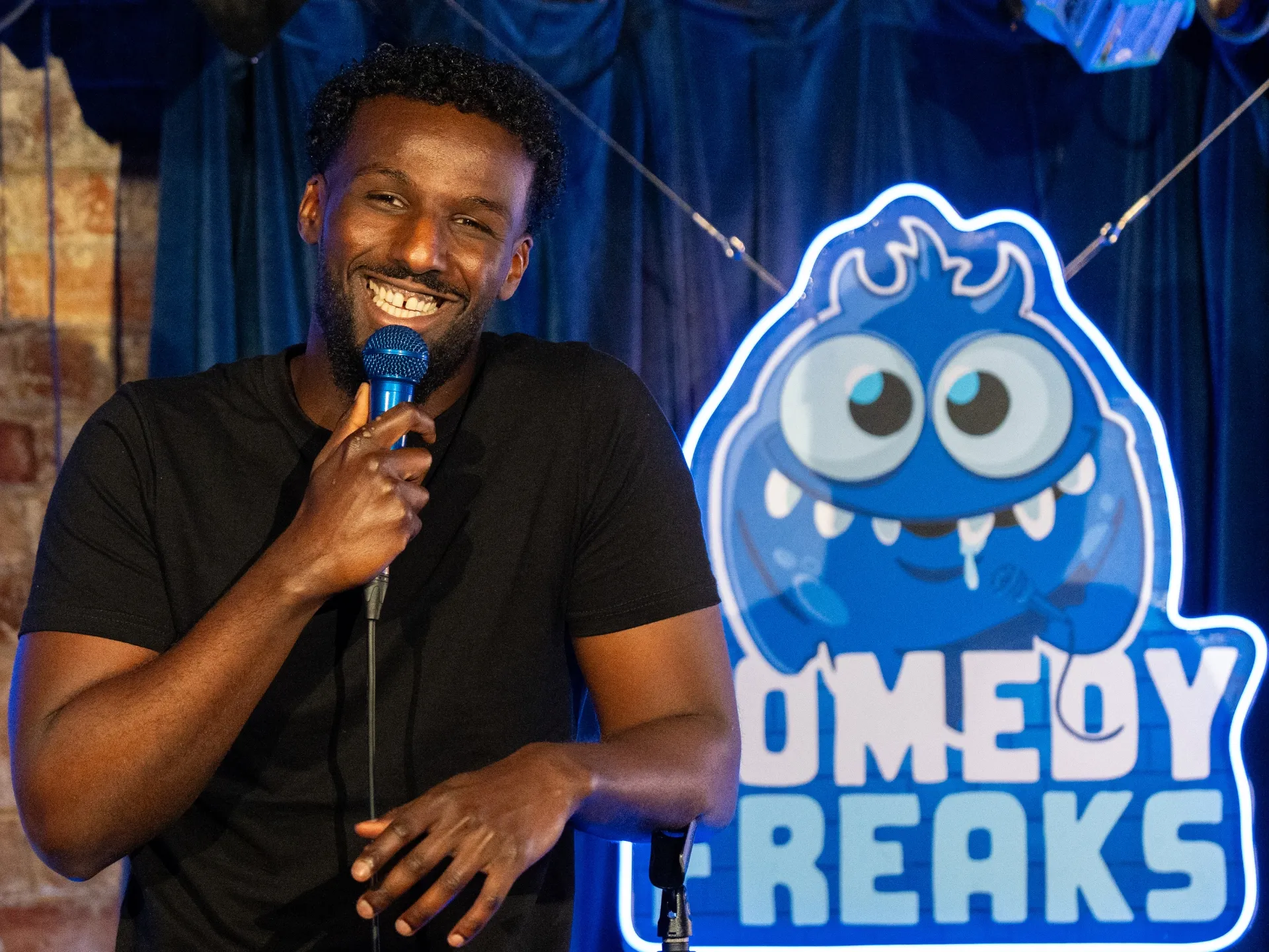 Comedian Bamph engaging the audience at a comedy show at Comedy Freaks, dressed in black casual wear, microphone in his left hand while gesturing with his right, illuminated by blue stage lighting with the glowing Comedy Freaks monster neon sign and brick architecture visible behind.