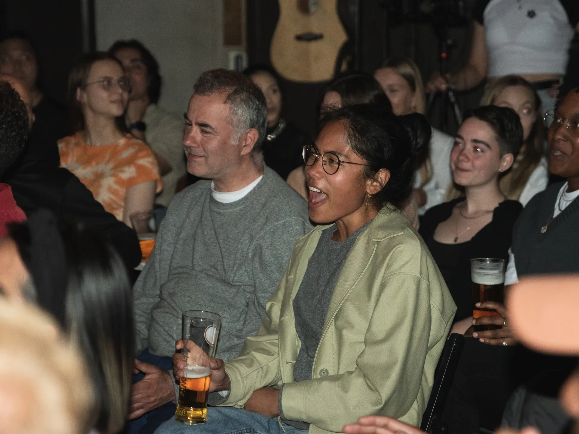 Packed audience at Comedy Freaks comedy show laughing and enjoying themselves, diverse group of patrons seated closely together holding drinks, woman in lime green jumper prominently visible in centre, warm ambient lighting creating an intimate house party atmosphere in the venue.