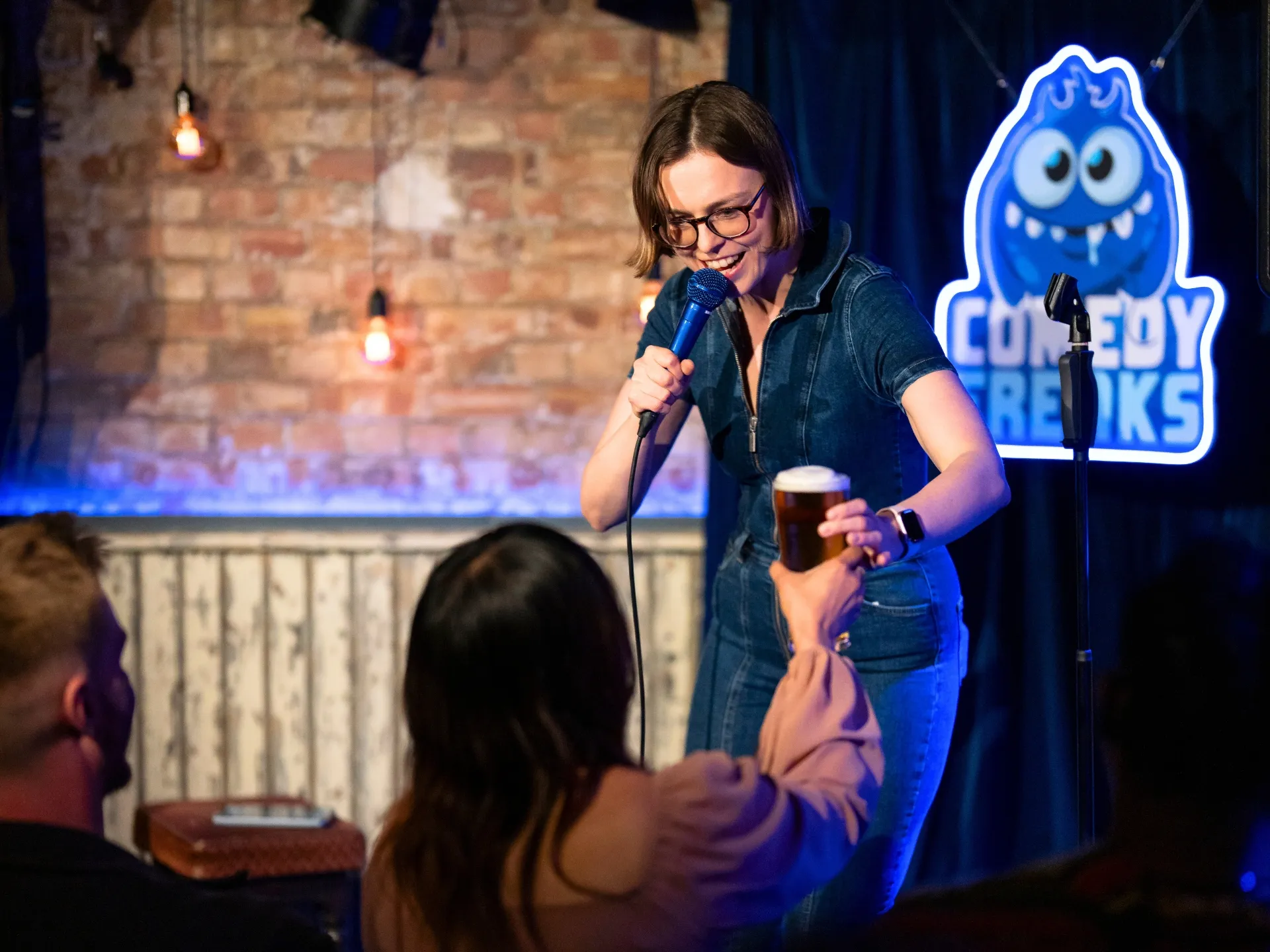 Comedian Katie Kamola delivering her set at a Comedy Freaks comedy show in London, wearing glasses and black denim jacket, casually holding a pint of beer while speaking into a microphone, with the vibrant blue Comedy Freaks monster logo and brick venue architecture behind her.