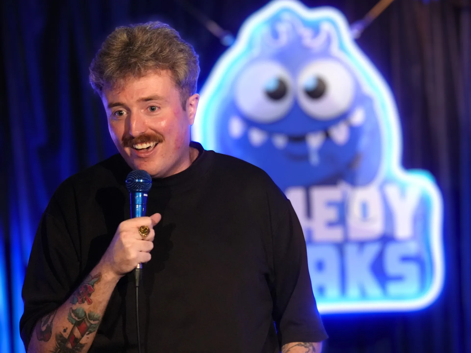 British comedian Jake Steers in a black crew neck t-shirt performing at a comedy show London venue at Comedy Freaks, gesturing naturally while speaking into a handheld microphone, with the iconic blue Comedy Freaks neon backdrop and dark purple curtains creating atmosphere behind him.