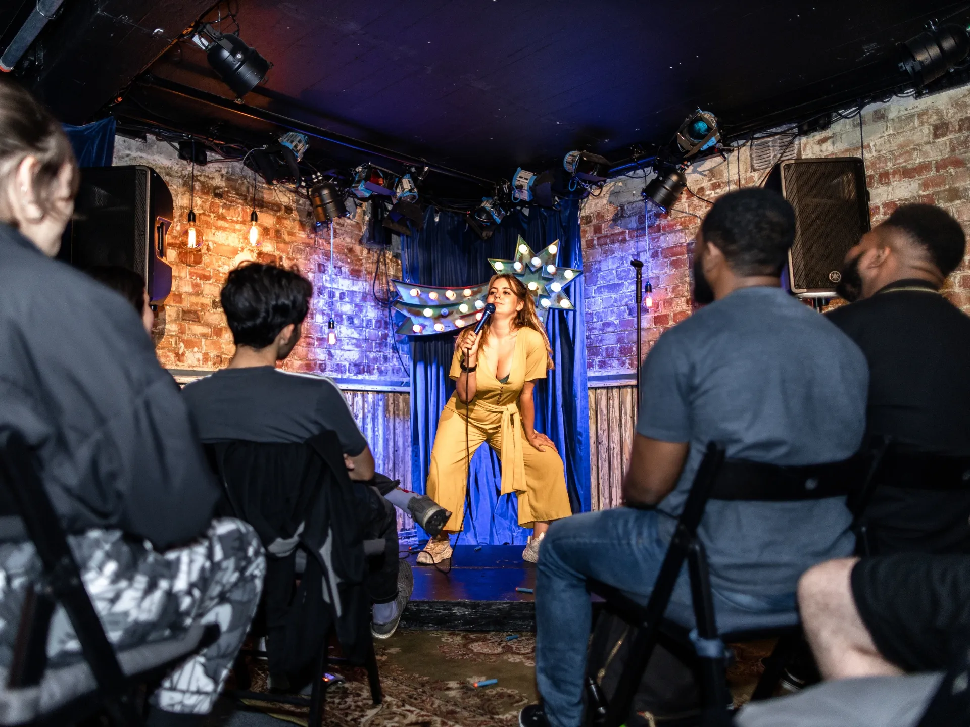 British Comedian Lou Barron mid-performance at a comedy show taking place at London comedy club - Comedy Freaks. Wearing a mustard yellow shirt and speaking into a microphone, with attentive audience members visible in the foreground and background, atmospheric stage lighting creating blue and purple hues throughout the venue.