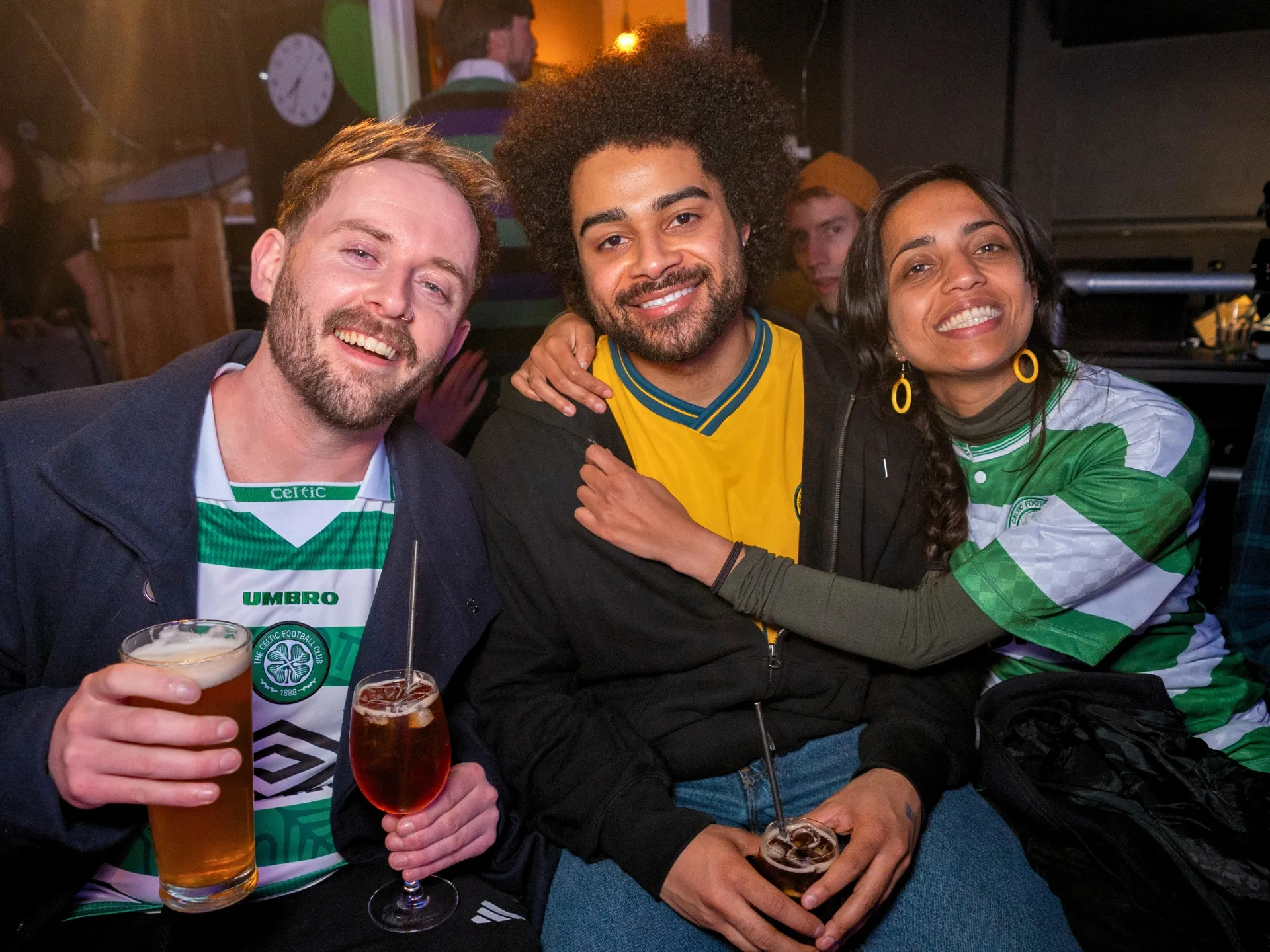 Three smiling audience members enjoying a Comedy Freaks comedy show in London, two men and one woman holding pints of beer, wearing green and white Celtic football jerseys, warm pub lighting creating a friendly atmosphere as they pose together during the interval with drinks in hand.