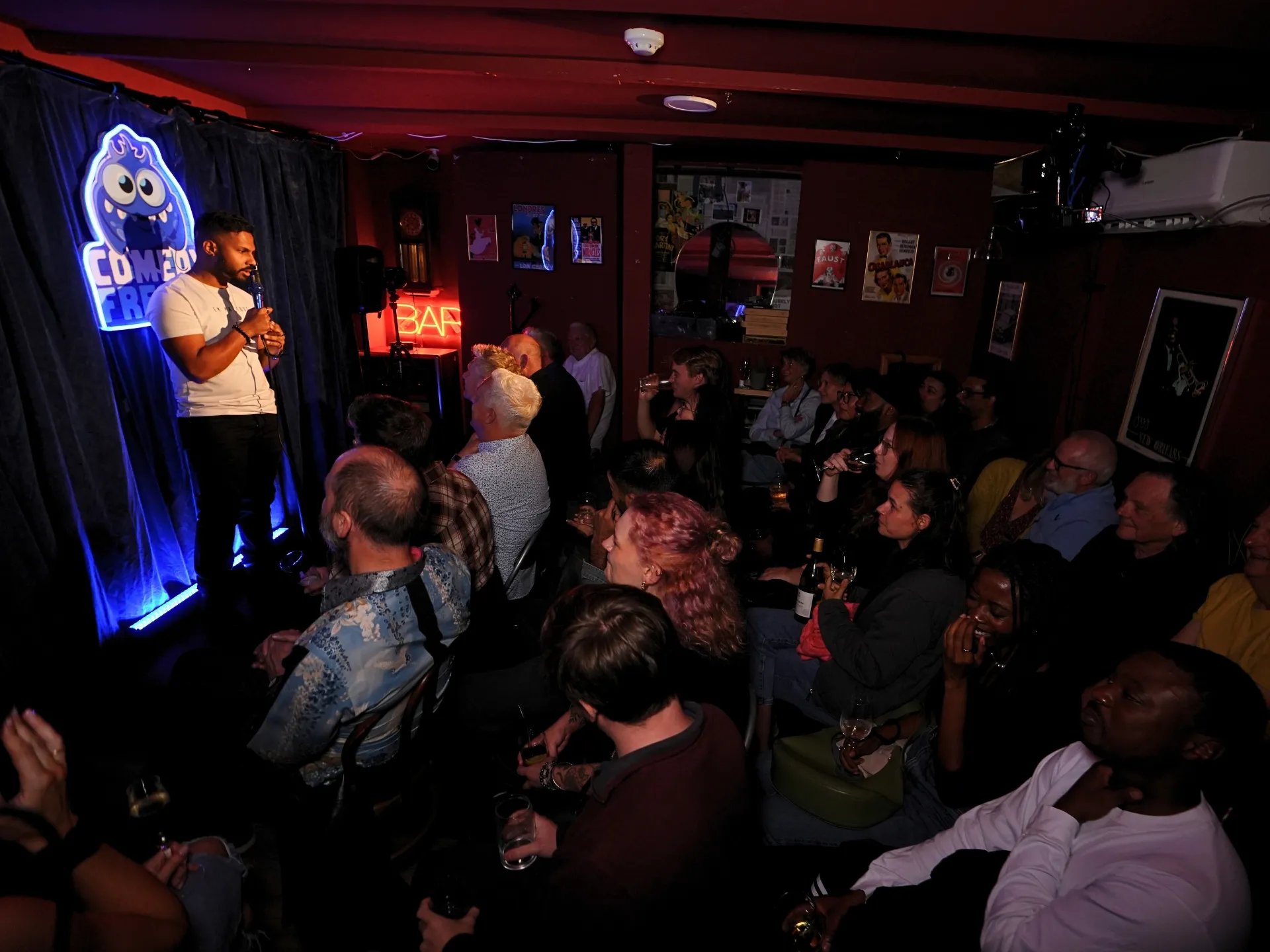 London comedian Raj Sivade commanding the stage at a Comedy Freaks London Comedy show, the best Camden comedy venue. The audience members seated with drinks watching intently beneath the glowing blue neon monster logo, intimate space buzzing with energy, red BAR sign and warm lighting throughout.