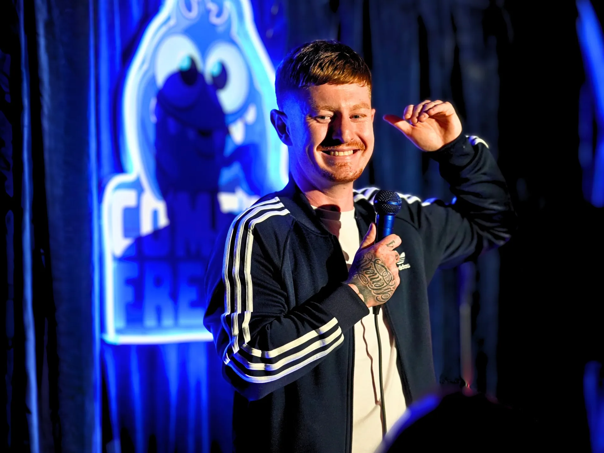  British comedian Tom Nestor on stage at a Comedy Freaks comedy show London event, wearing a sporty black Adidas track top, short ginger hair catching the blue stage lighting, holding the microphone close while smiling, the iconic neon Comedy Freaks logo glowing brightly behind him.