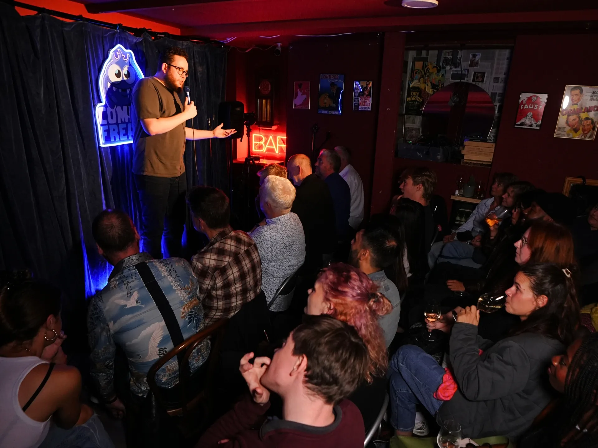 UK comedian Timoteo Kiss Freitas performing at a Comedy Freaks comedy show at the London comedy club, wearing a brown shirt and gesturing expressively with open hands while addressing the packed audience, blue neon logo and red bar sign glowing in the background, crowd visible in the foreground.