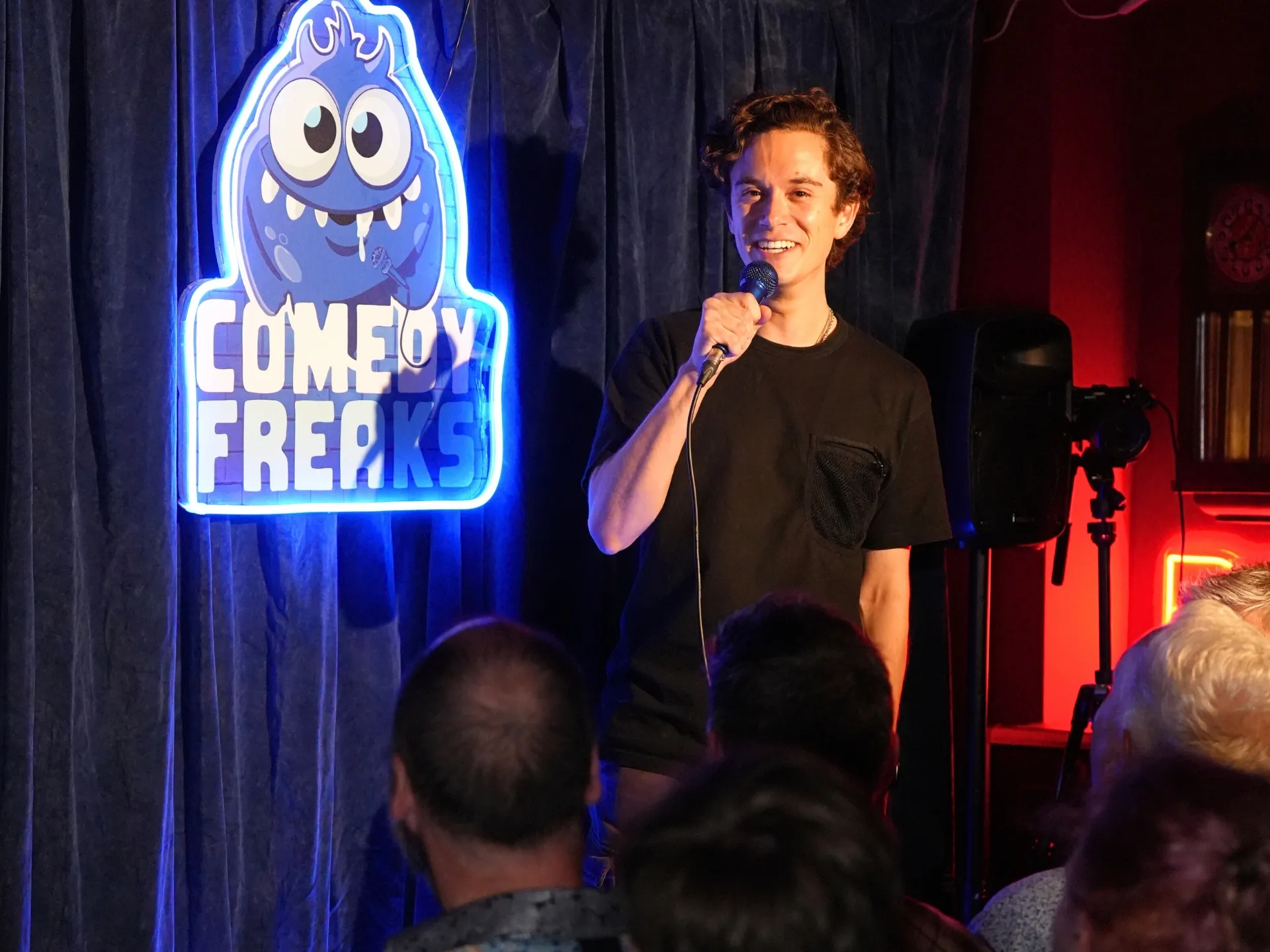UK comedian Jomi Cruz mid-set at a London comedy show at Comedy Freaks. He has dark curly hair and relaxed expression as he speaks into the microphone, wearing a simple black tee, the iconic blue neon monster logo and red ambient lighting creating a vibrant stage backdrop.