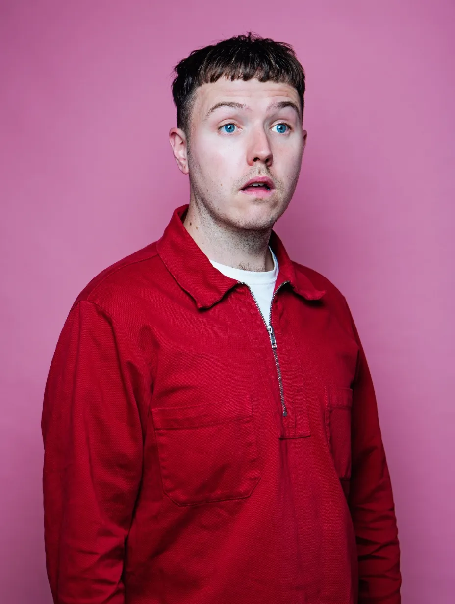 Comedian Adam Flood photographed against hot pink wearing a red zip-up jacket over white. The BBC New Comedy Award nominee has striking blue eyes and a playfully surprised expression. Catch him at Comedy Freaks for top London comedy shows.
