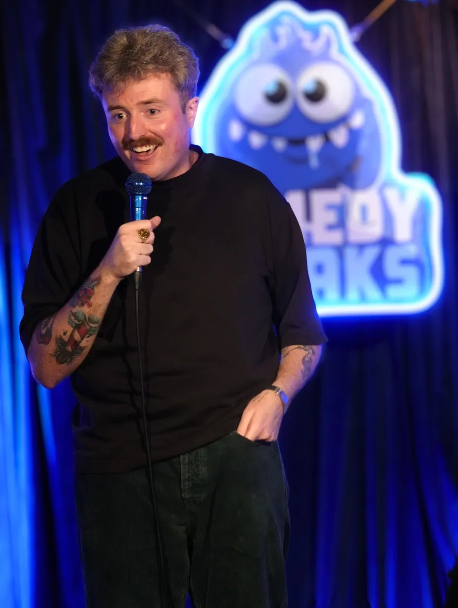 Stand-up comedian Jake Steers mid-performance at London comedy club - Comedy Freaks. He's smiling and holding a microphone. He is wearing a black t-shirt with visible tattoos on both forearms, and the blue Comedy Freaks logo is lit up on the wall behind him.