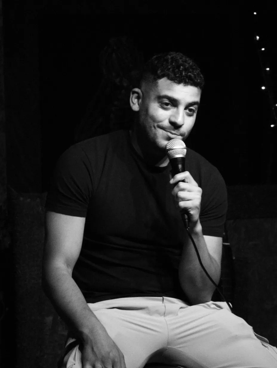 Ashley Bay, a London comedian performing stand-up comedy at Comedy Freaks, seated on stage in a black t-shirt and light trousers, holding a microphone close to his face. He has short curly hair and a subtle smile, photographed in black and white.