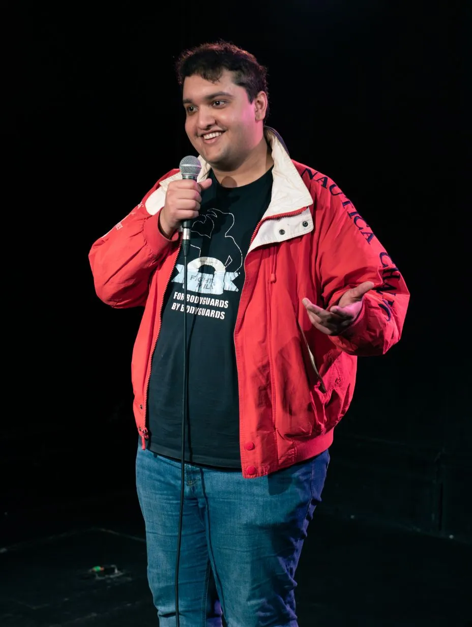 Image description: Rez Bhatt performing on a dark stage. Full-body performance shot. He wears a red Nautica jacket over a black graphic t-shirt (reads "For Bodyguards By Bodyguards") and blue jeans. Holding a microphone stand with his right hand, right arm raised. Smiling warmly at the audience. Warm pink-toned overhead stage lighting. Dark background. 