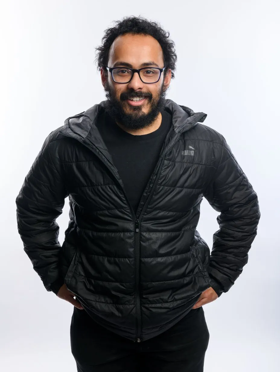 Professional studio headshot of Ridwan Hussain. He has curly dark hair, a full beard, and wears blue rectangular-framed glasses. He is dressed in a black hooded Puma puffer jacket over a black t-shirt. He stands with both hands on hips, smiling broadly and directly at the camera. The background is a clean, pale grey/white studio background.