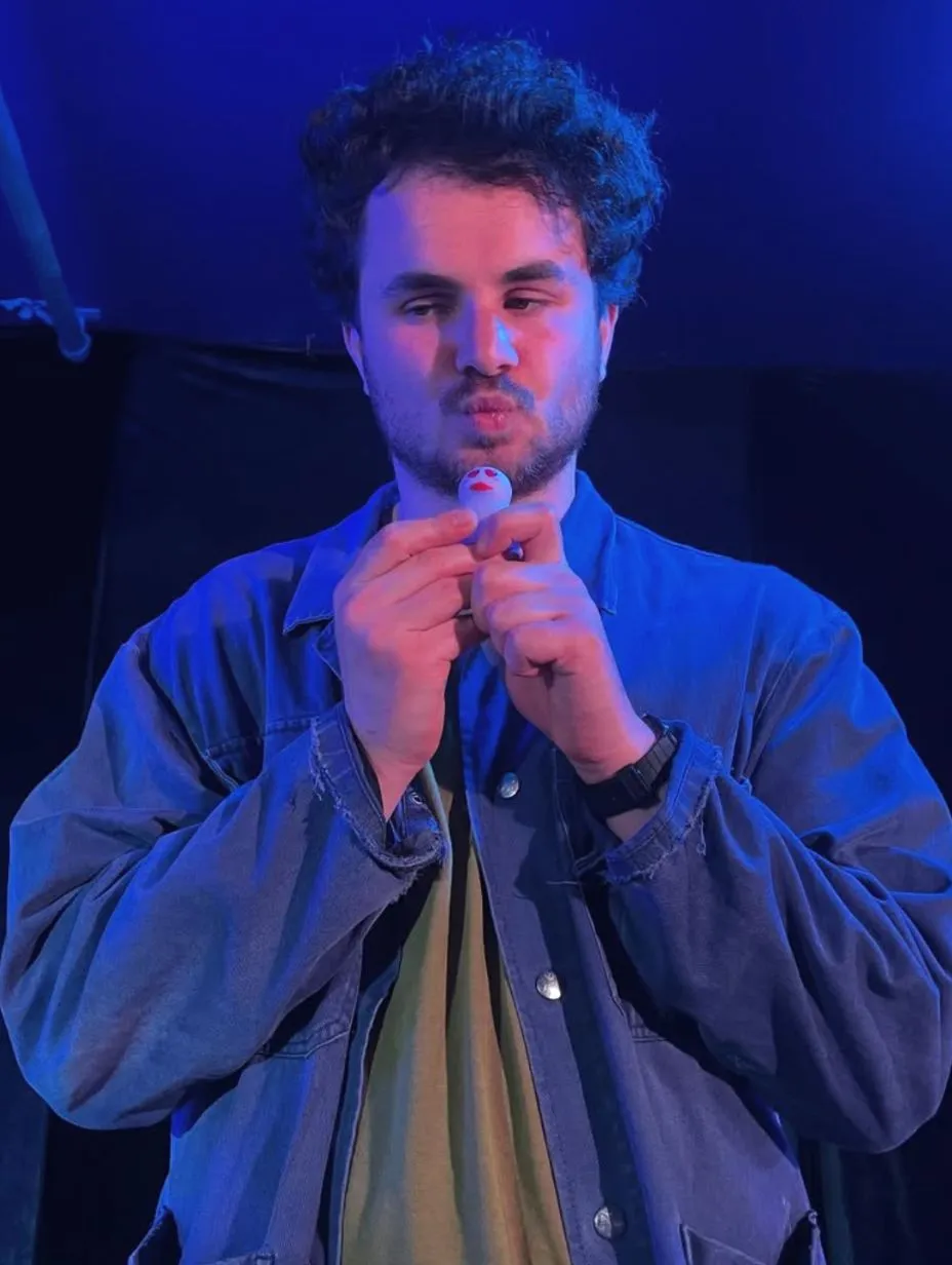 London comedian George Lando mid-performance, holding a microphone to pursed lips with both hands and looking sideways. He has curly dark hair, light stubble, and wears a blue denim jacket over an olive t-shirt.