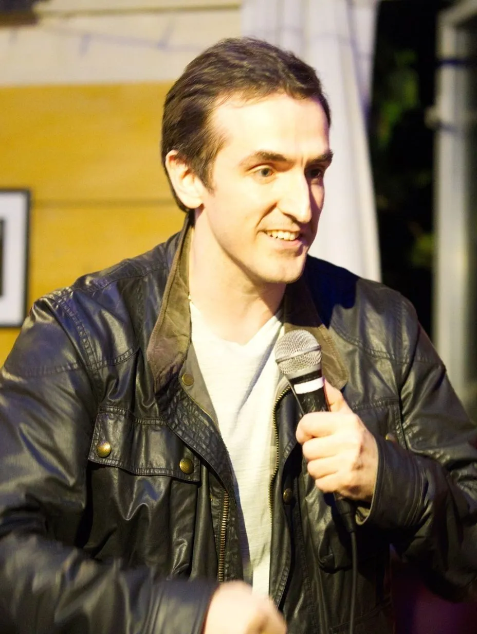 Live performance shot, likely outdoors or in an informal venue. Cathal O'Riordan has short, neatly combed dark brown hair, light skin, and prominent cheekbones. He is smiling broadly and looking slightly to the right of camera, with a natural, engaged expression mid-set. He holds a handheld microphone in his right hand at about chest height. He wears a black leather jacket (with front flap pockets and metal snap buttons) over a white/light grey crew-neck t-shirt. The background is warm and naturally lit: yellow-toned wooden panelling to the left, a white curtain/backdrop upper right, and soft ambient light — no heavy stage wash. A partial audience member is visible at the bottom right. Shot is cropped from roughly the waist upward.