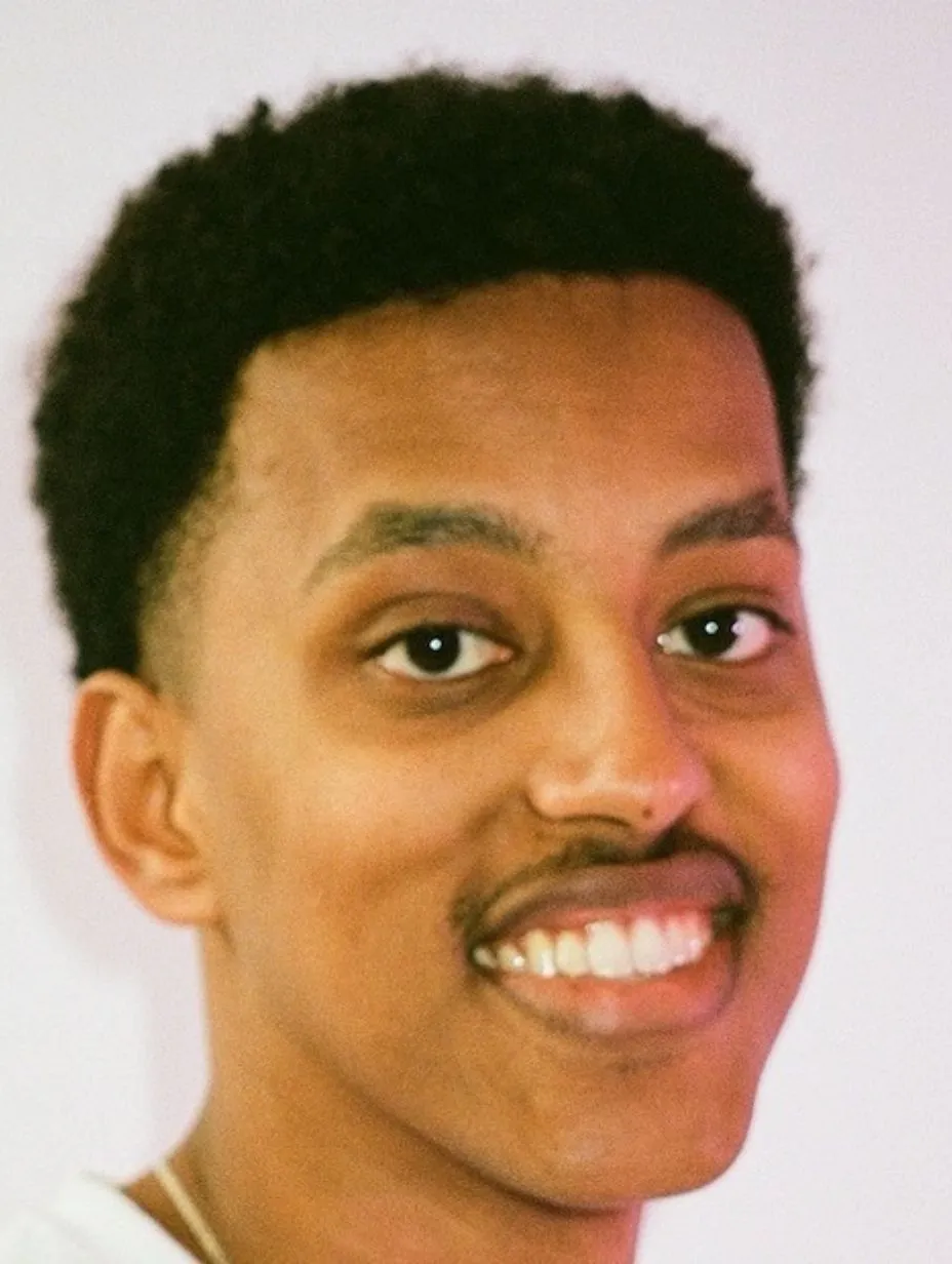 A close-cropped headshot portrait. Ramy Shafi has short, natural coily/textured black hair, a thin moustache, warm medium-brown skin tone, and a broad, bright smile showing his teeth. He is looking slightly to the right of camera. He wears a white or off-white top, and a thin gold/delicate chain necklace is just visible at the base of the frame. The background is plain pale grey/lavender-white. Shot is cropped from roughly the upper chest/collar upward — primarily a face and head portrait.