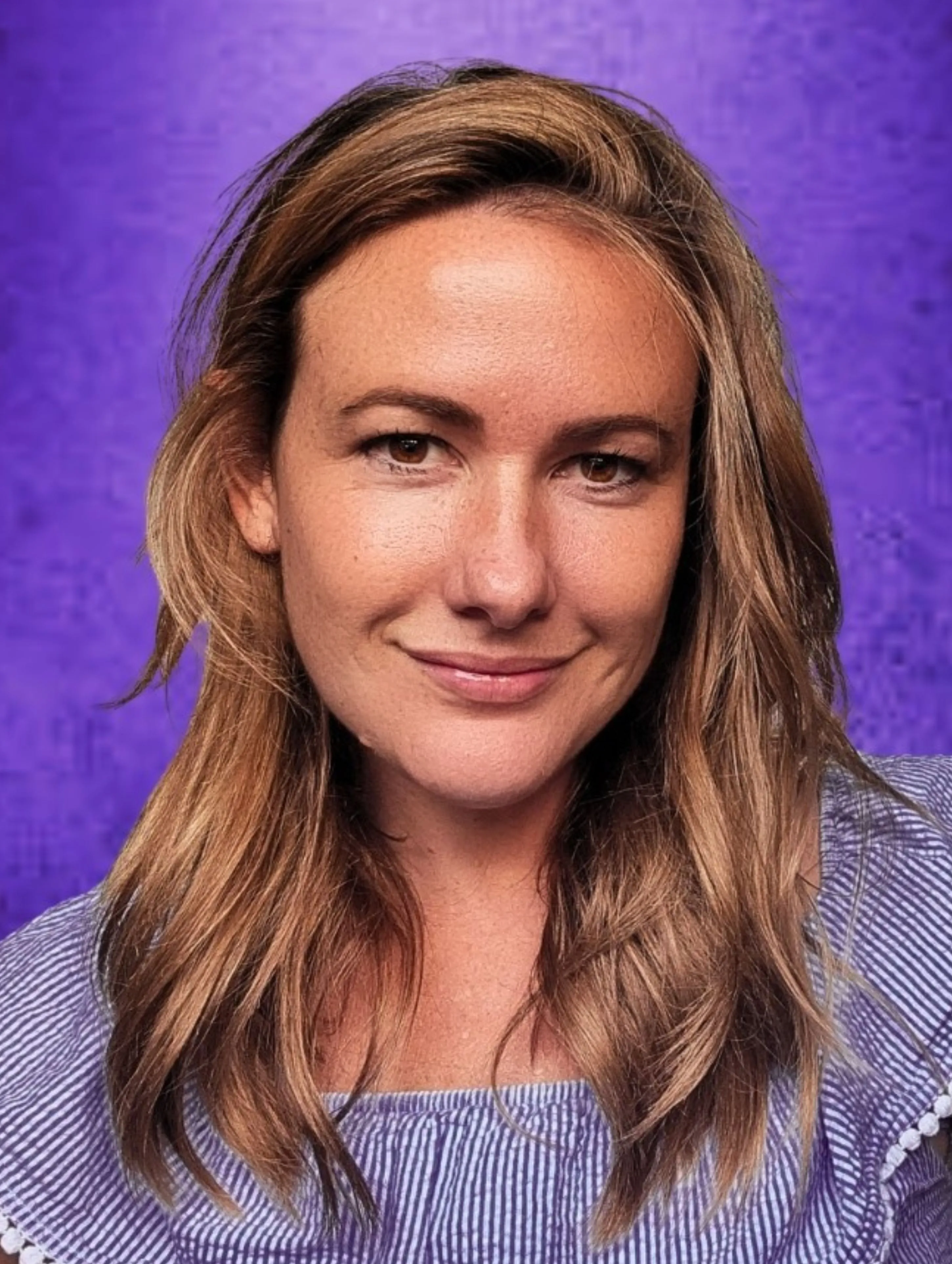 Comedian Laura McMahon in a professional studio headshot, smiling warmly at the camera. Medium-length wavy brown hair with lighter highlights, wearing a light blue fine-striped top. Vivid purple studio backdrop