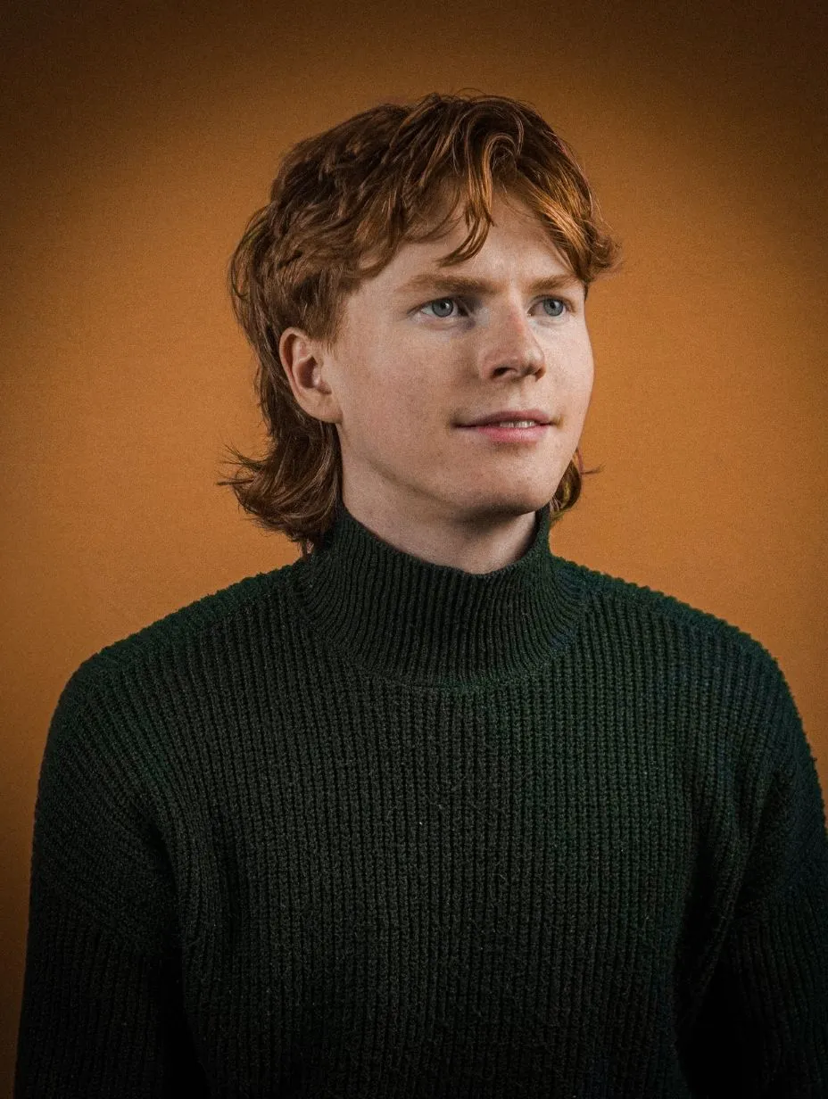 Comedian Lucas Jefcoate in a professional studio headshot. Medium-length wavy auburn hair, blue eyes, slight smile looking off-camera. Dark forest green ribbed turtleneck jumper, warm amber studio backdrop.