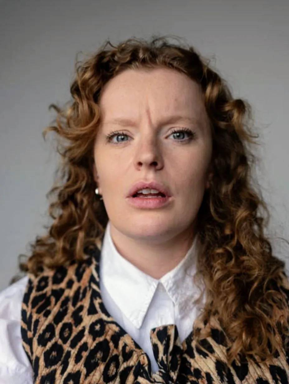 Comedian Lauren Soley in a professional studio headshot. Curly auburn hair, blue-grey eyes, slight open-mouth expression. White collared shirt under a leopard print vest, small stud earring.