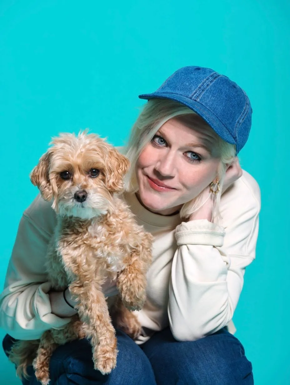 Comedian Lulu Popplewell in a professional studio headshot, smiling and holding a small curly sandy-coloured dog. Blue denim baseball cap, cream sweatshirt, gold hoop earrings. Vivid turquoise studio backdrop.