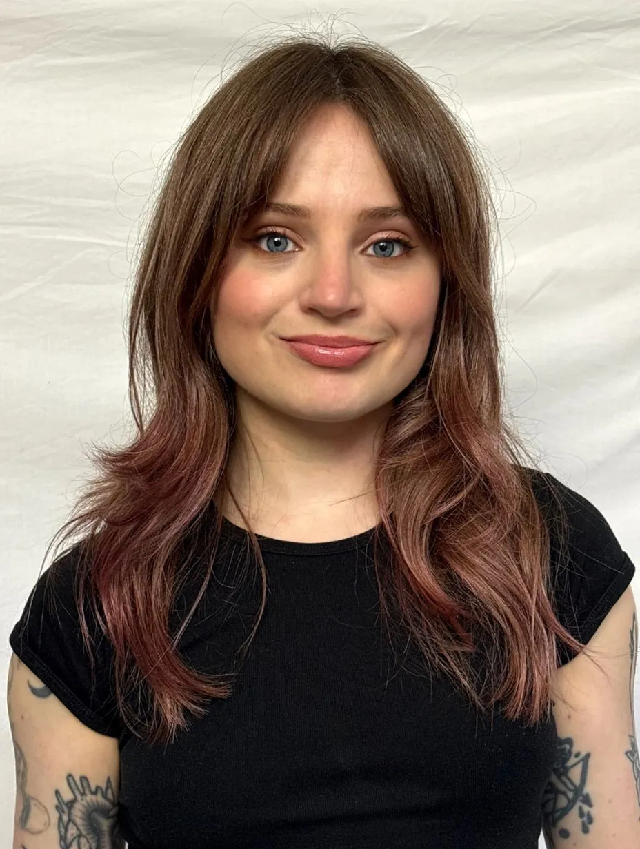 Comedian Kat Baker in an informal portrait headshot. Dark brown hair with reddish ends and curtain fringe, blue-grey eyes, warm slight smile. Black fitted t-shirt, tattoos on both forearms. White fabric backdrop.