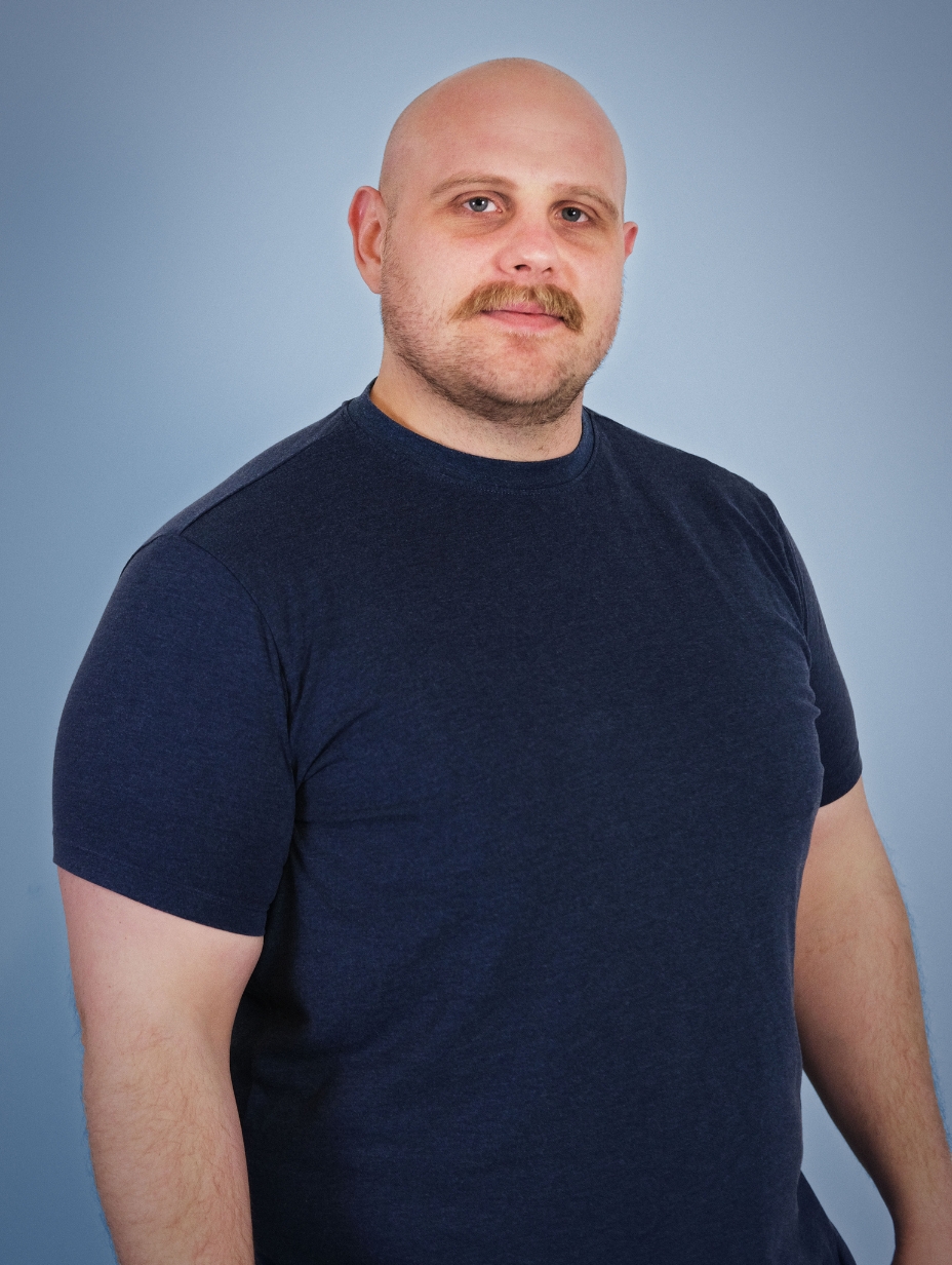 Comedian Hamish Shepherd in a professional studio headshot. Shaved head, light moustache and jaw stubble, calm expression looking slightly off-camera. Dark navy blue t-shirt, soft blue-grey gradient studio backdrop. 