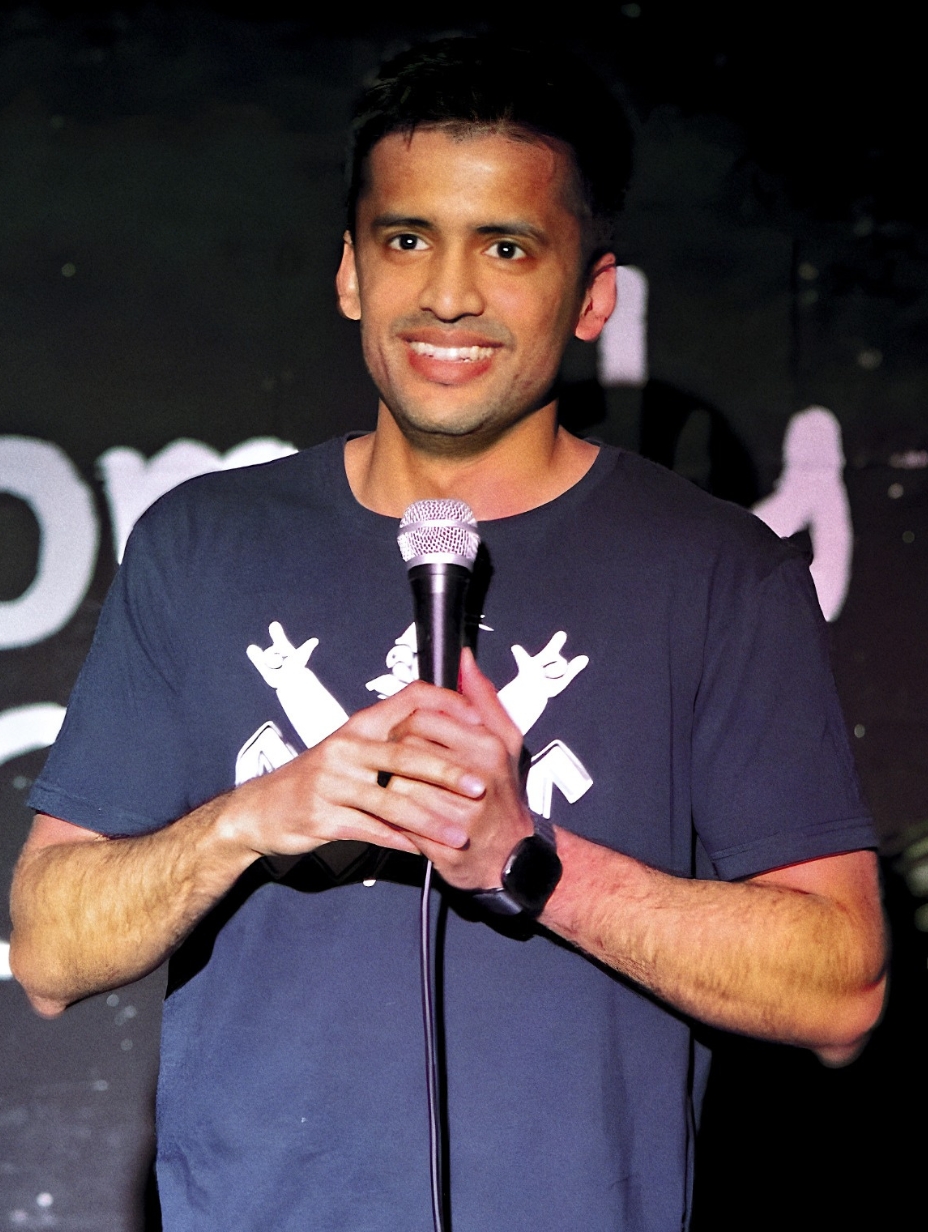 Comedian Aayush Rathi performing live on stage, smiling broadly while holding a microphone with both hands. Dark navy blue graphic t-shirt, dark event backdrop with partial text branding, warm stage lighting. 