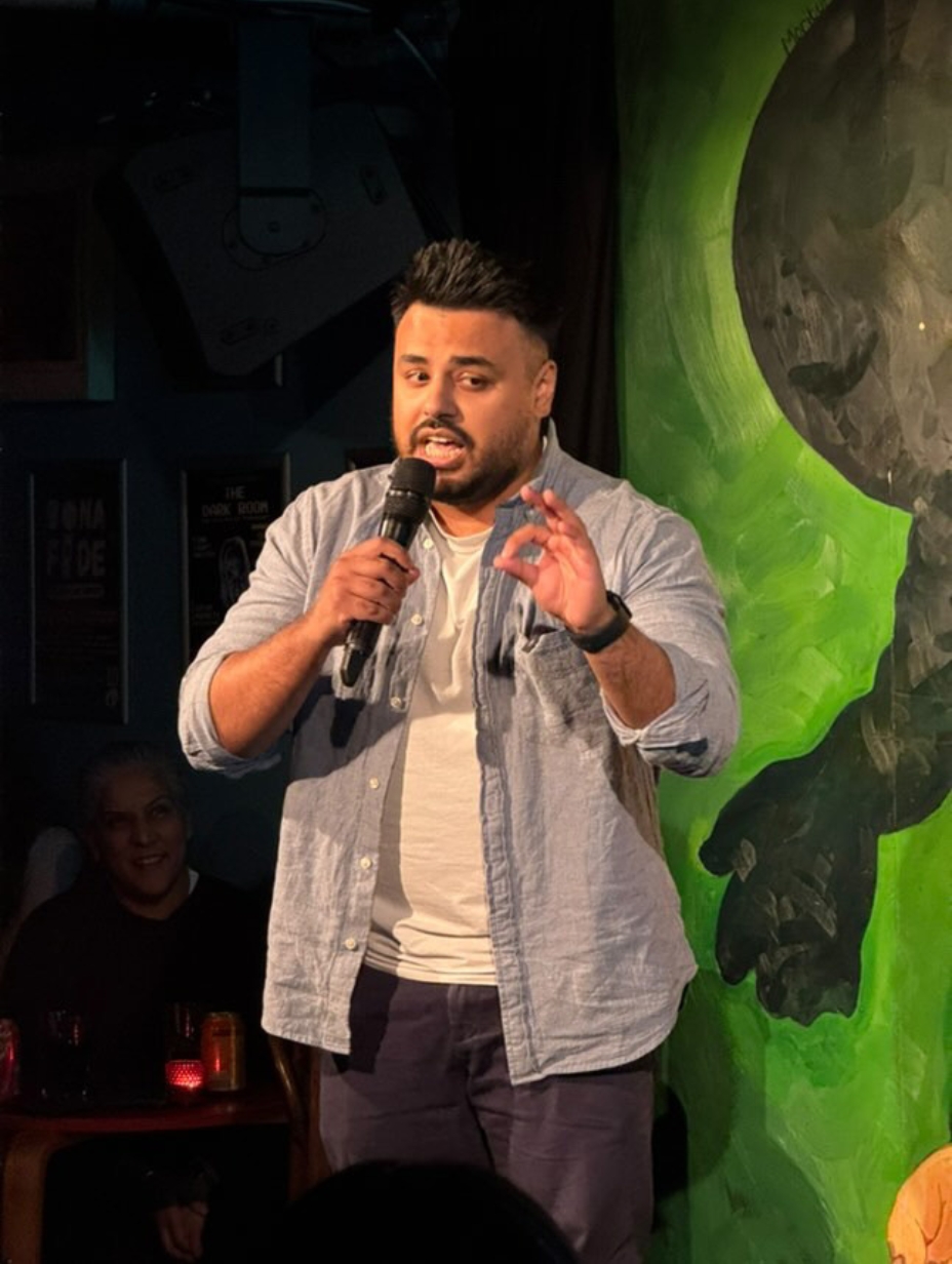 Comedian Nayim Ali performing live on stage, holding a microphone with one hand and gesturing with the other. Light blue open shirt over white t-shirt, dark trousers. Dark background with a vivid green painted mural. 
