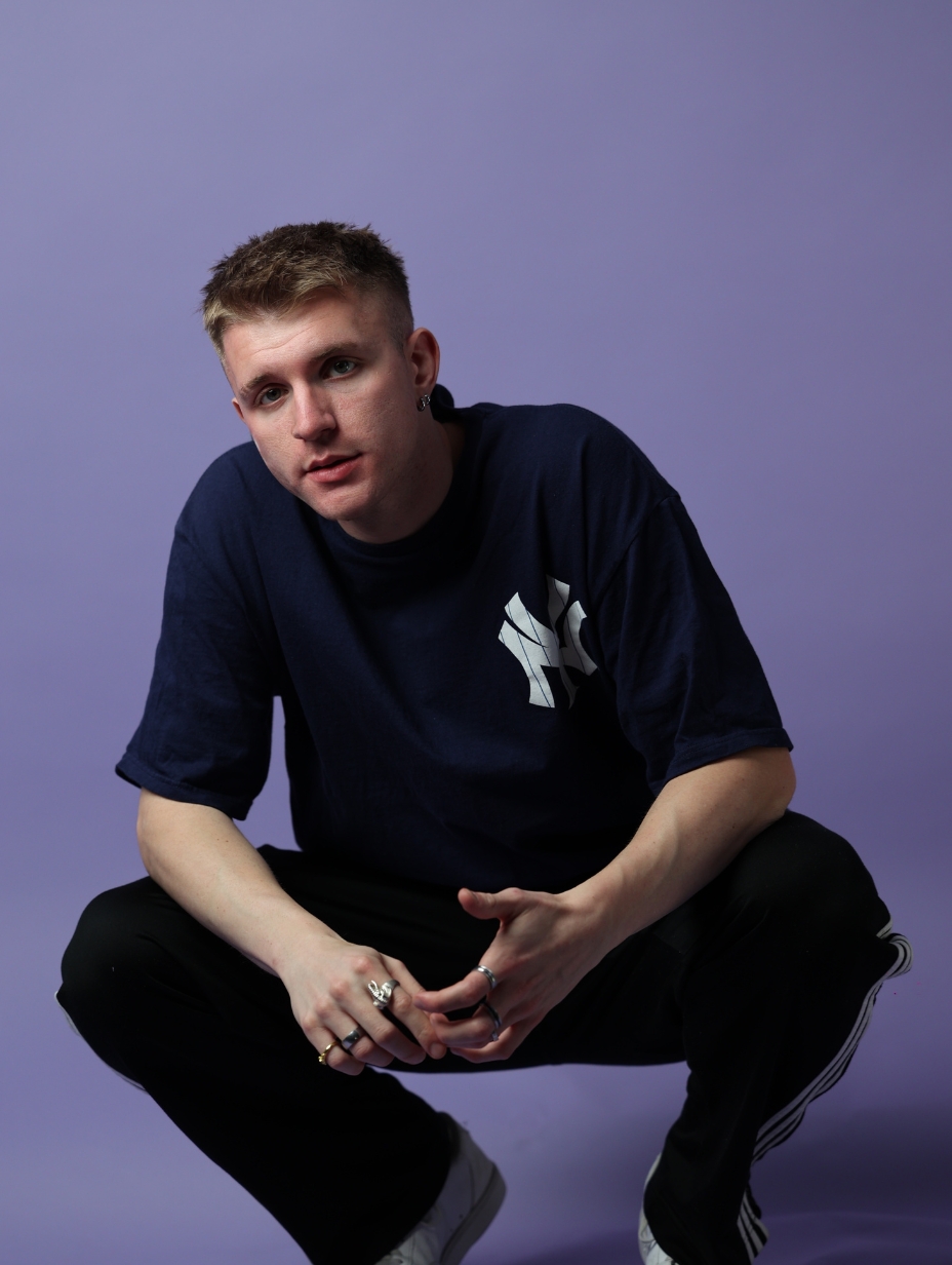 Comedian Zak Ranger in a studio headshot, squatting with hands clasped. Short sandy hair, small stud earring, multiple rings. Dark navy NY Yankees t-shirt, black Adidas tracksuit bottoms. Soft lavender backdrop. 