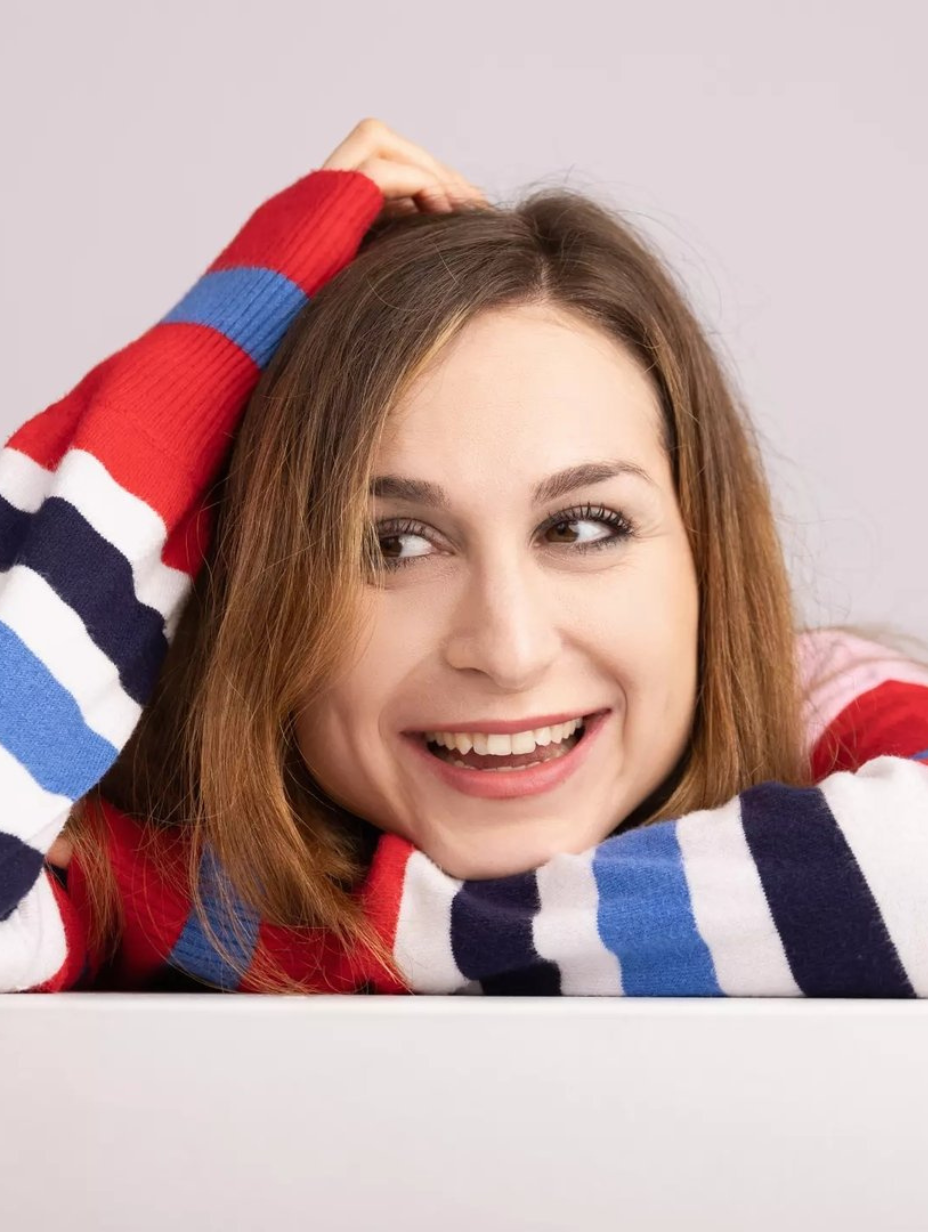 Comedian Hannah Lloyd-Davies in a studio headshot, resting her arms on a white surface and smiling sideways. Shoulder-length brown hair, bold red white blue and navy striped jumper. Soft grey studio backdrop.