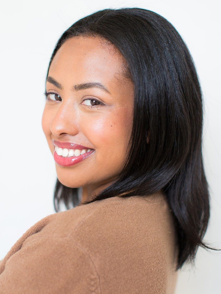 Comedian Arie David in a studio headshot, looking over her shoulder at the camera with a warm smile. Long straight black hair, light brown eyes, nude-pink lip colour. Camel brown coat. White studio backdrop.