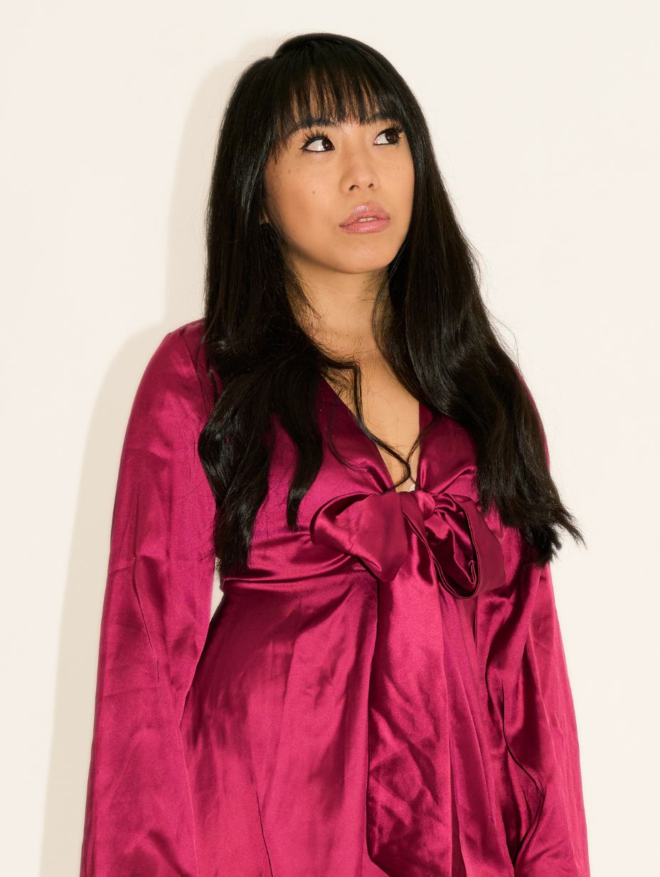 Comedian Ginnia Cheng in a professional headshot, looking upward with a wry expression. Long straight black hair with fringe, wearing a deep magenta satin wrap dress with bow detail. Off-white studio wall backdrop.