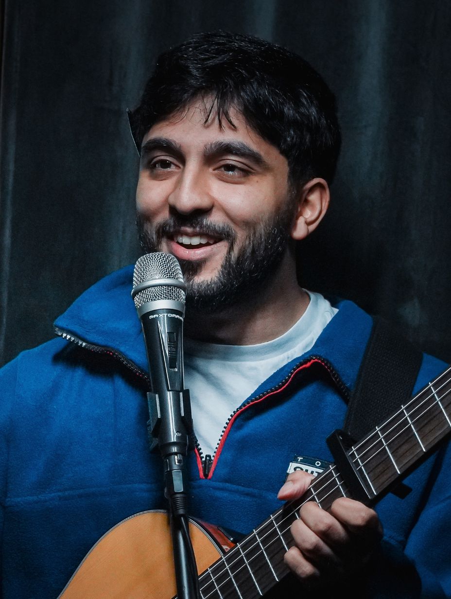 Comedian Paras Patel performing on stage, smiling while holding an acoustic guitar at a microphone stand. Short dark hair, dark beard, blue zip-up fleece over white t-shirt. Dark curtain backdrop, warm stage lighting. 