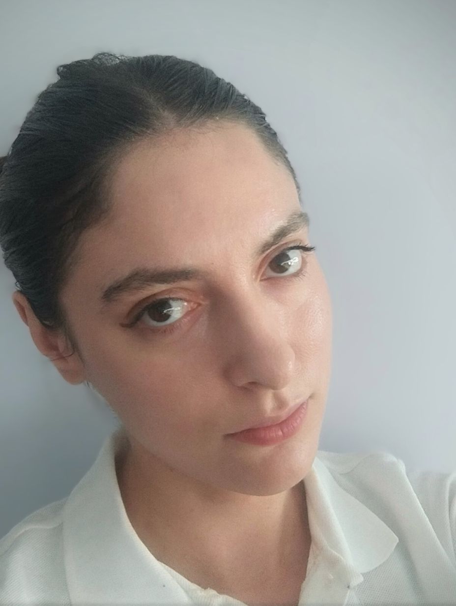 London comedian Francesca Rainey in an informal portrait headshot. Dark hair neatly pulled back in a bun, dark eyes, calm direct expression. White ruffled-collar top. Plain light blue-grey wall backdrop. 