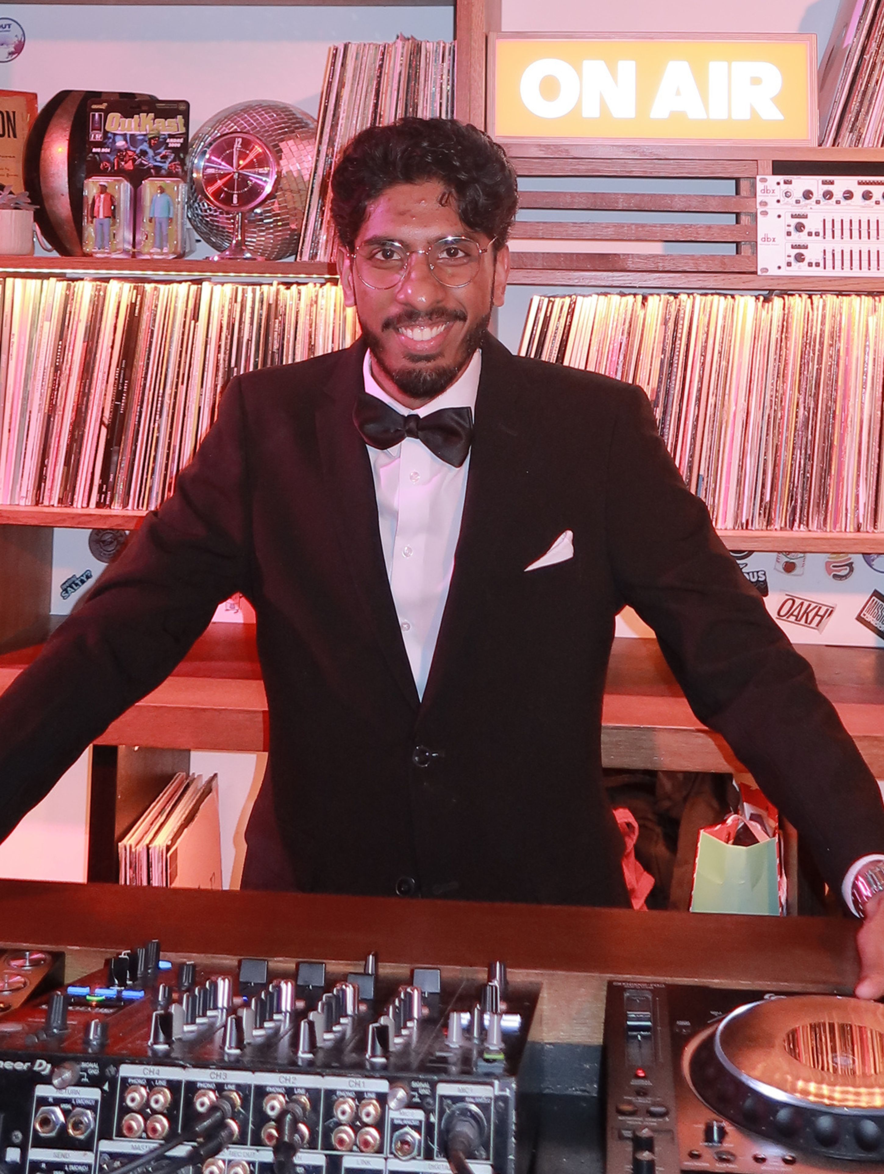 London comedian Sobin ST in a black tuxedo with bow tie and round glasses, smiling confidently behind DJ decks surrounded by vinyl records at a Comedy Freaks comedy club London event, with an illuminated ON AIR sign glowing amber in the background.